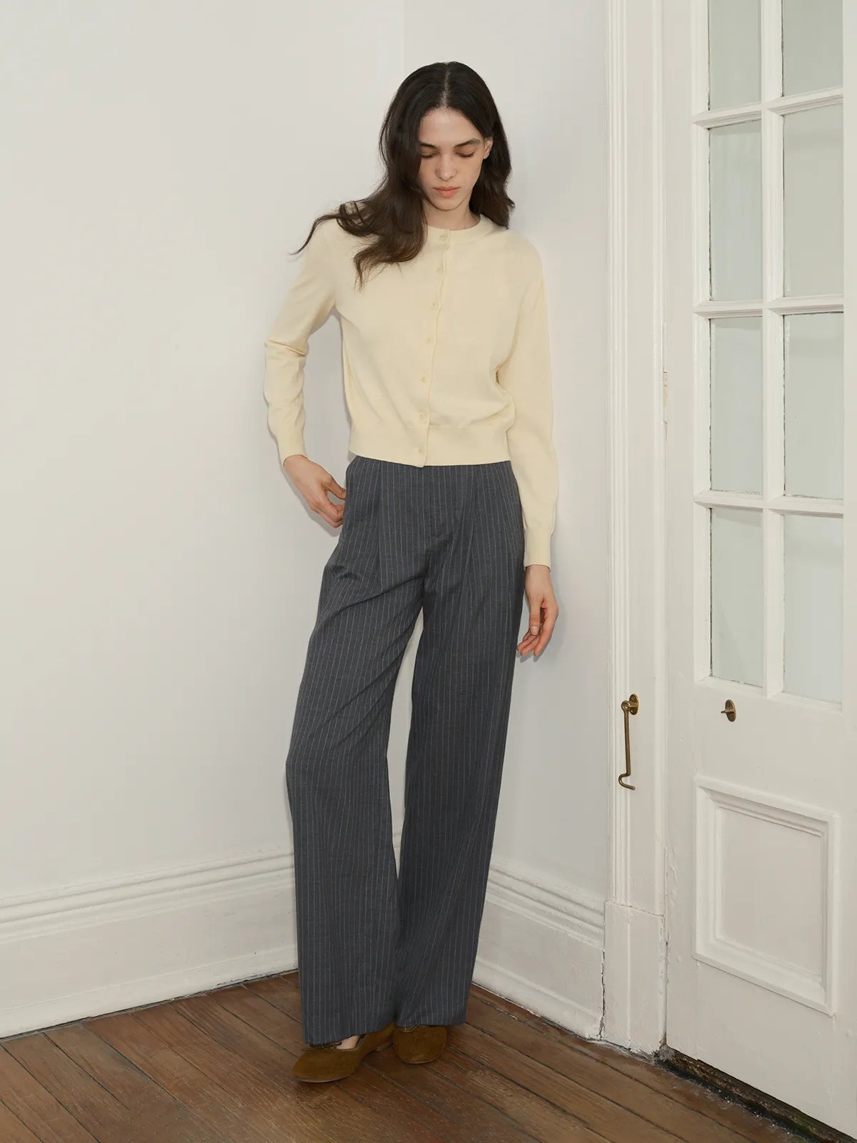Model wearing BINX slightly relaxed cardigan in ivory with grey pinstripe trousers, standing by white door