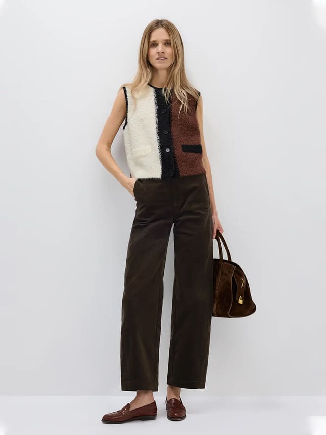 Model wears a sleeveless Brown/White/Black colorblock knitted vest with front buttons and a pocket, layered over a white blouse and black trousers.