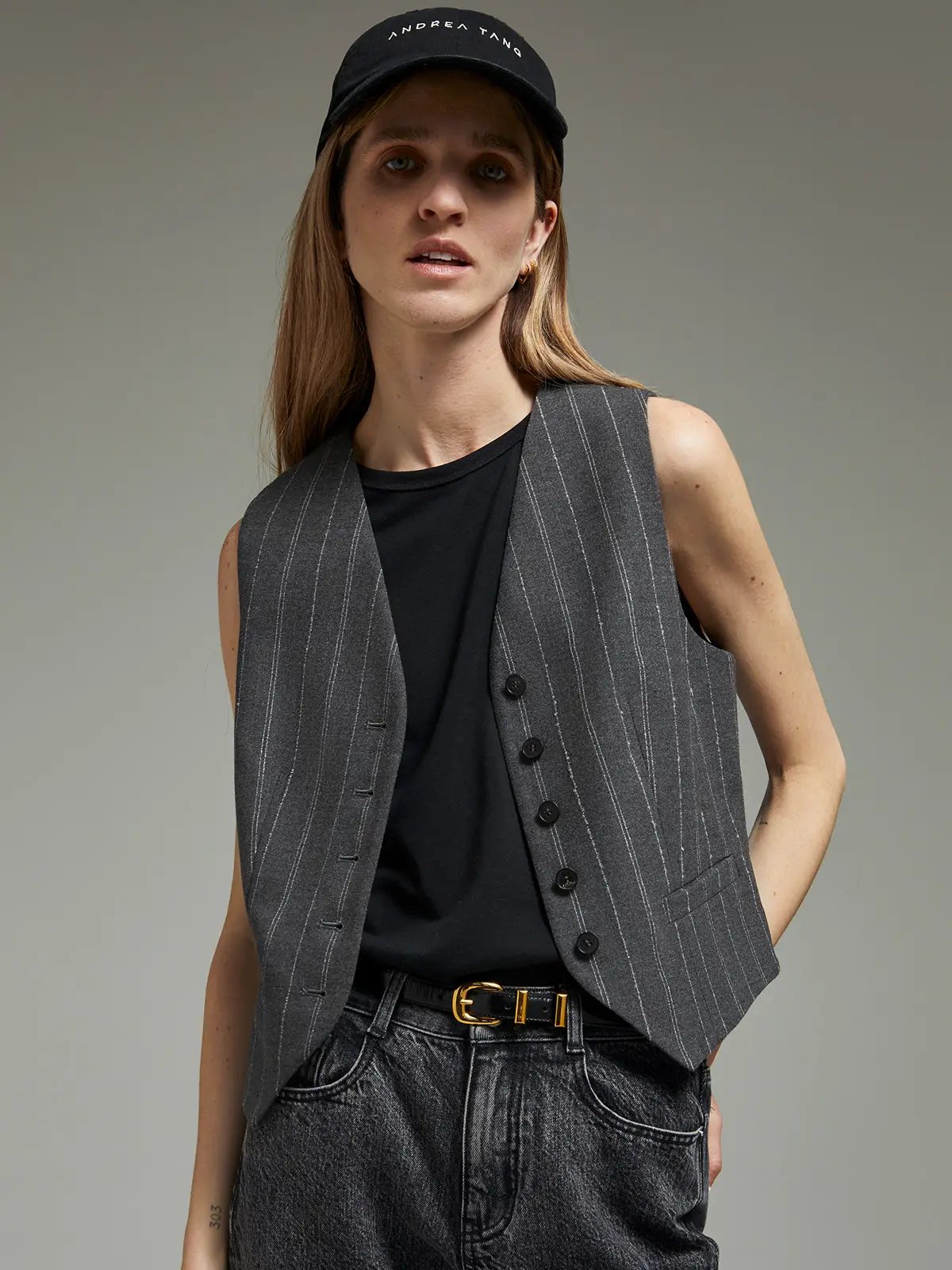 Grey pinstripe wool vest with five buttons, worn over a tee, dark jeans, and a cap.