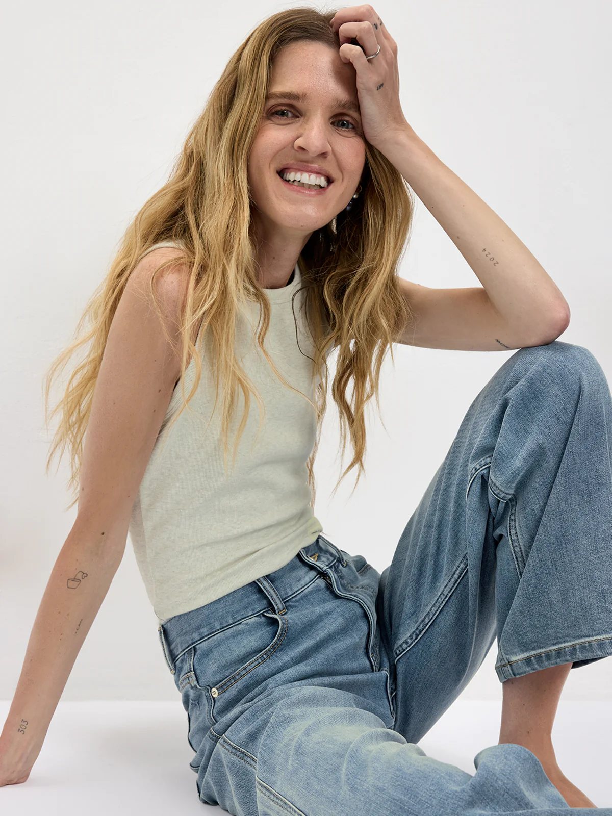 Model seated wearing a Light green fitted tank top and wide-leg jeans, hand resting on head.