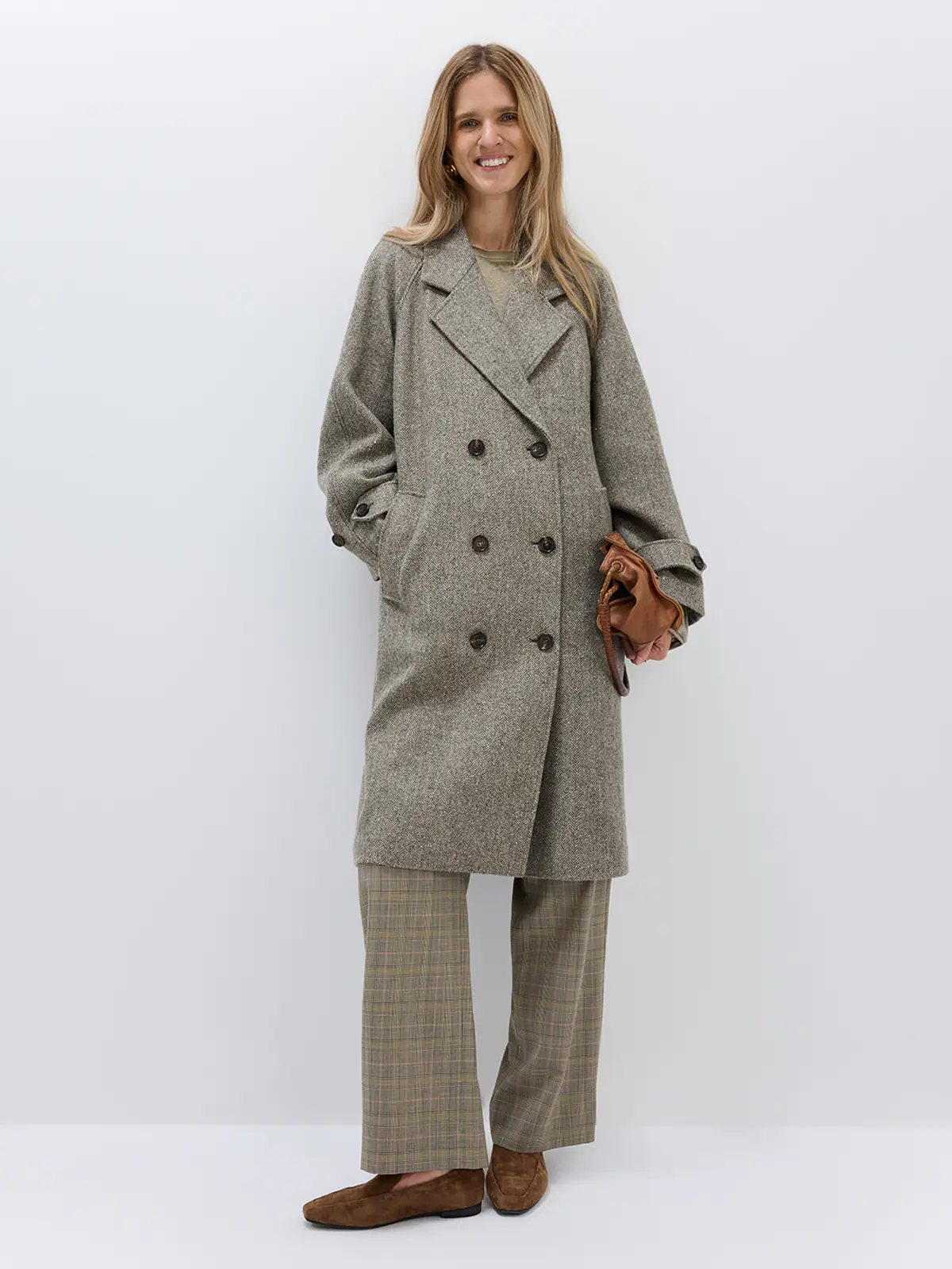Front view of a model wearing a brown herringbone double-breasted coat with wide lapels and six dark buttons, hands in pockets.