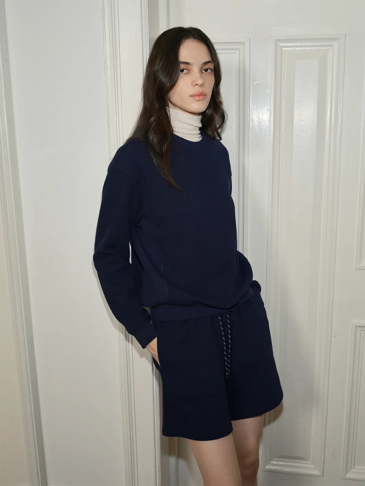 Model in navy SOREN sweatshirt and shorts leaning against white paneled door, hands in pockets