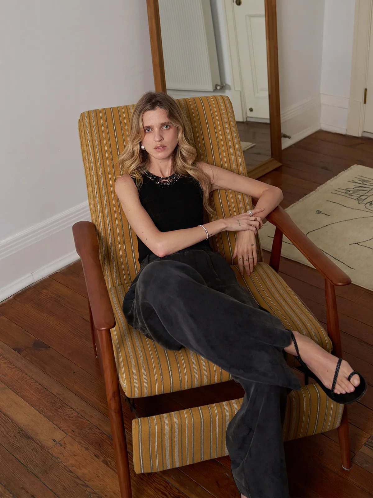 Model seated in vintage armchair wearing black MEADOW Textured Knit Tank Top with beaded neckline detail and dark velvet trousers