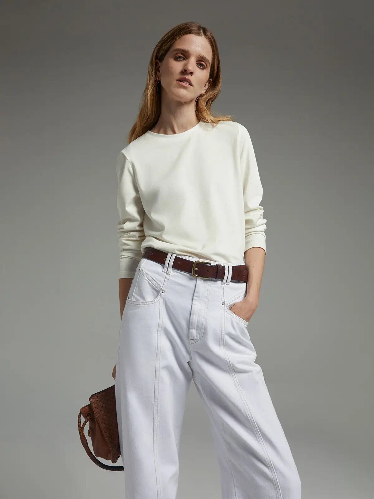 Model wearing a white long-sleeve crewneck T-shirt with rolled sleeves, tucked into white wide-leg jeans with a belt, holding a small bag.