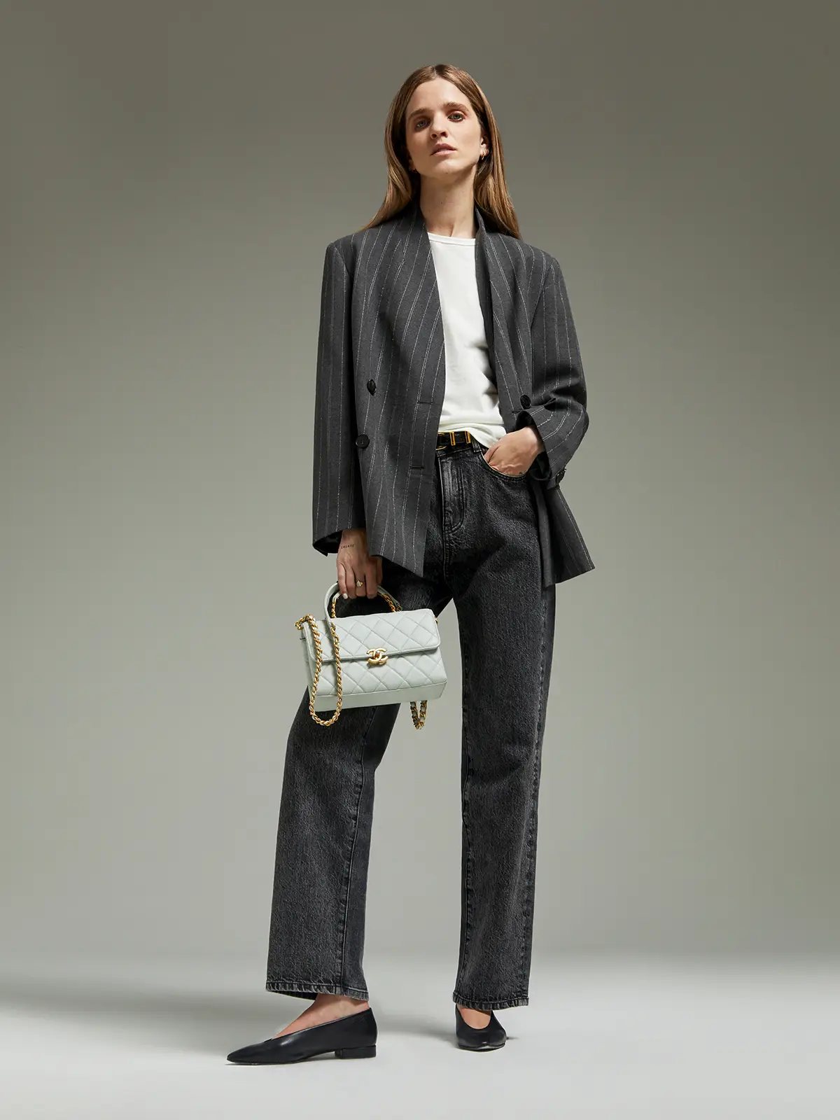 Full front view of model in gray pinstripe blazer holding a light handbag, paired with dark jeans and flats.
