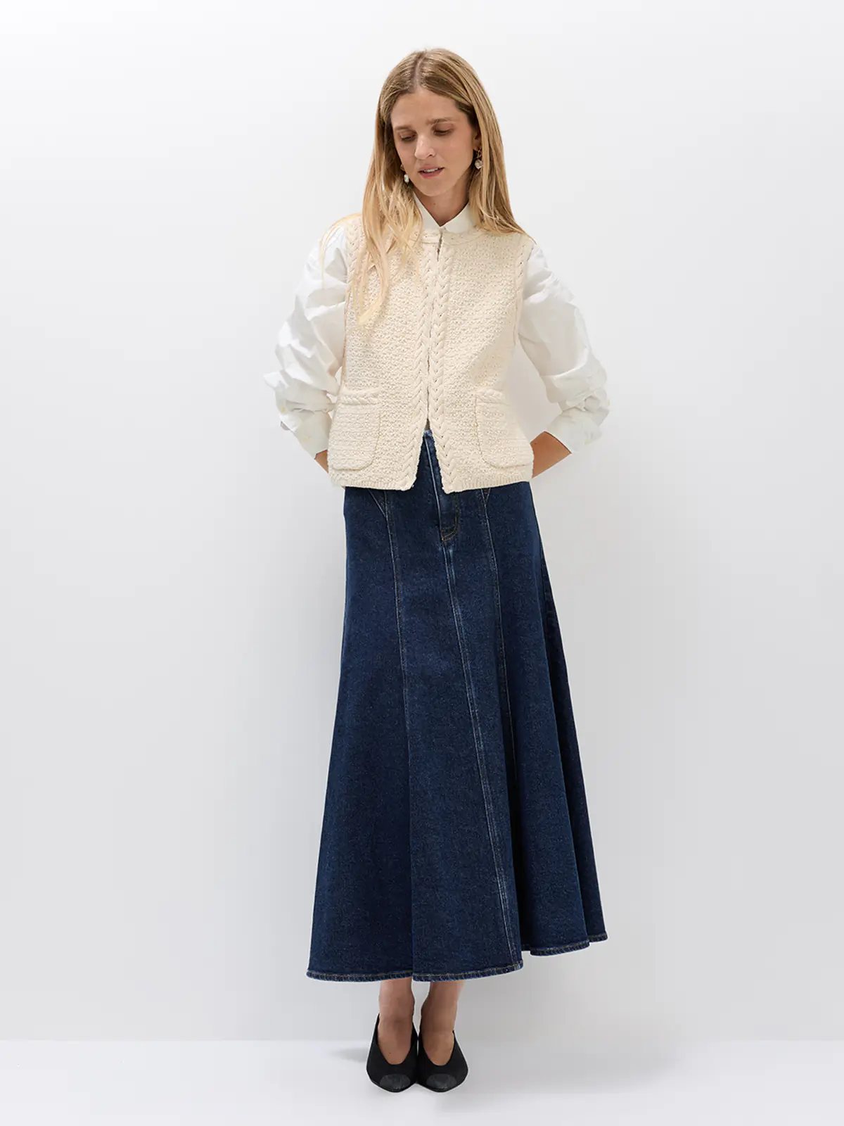 Off-white sleeveless crochet vest with cable detailing and front pockets, worn over a white blouse with a dark denim skirt.