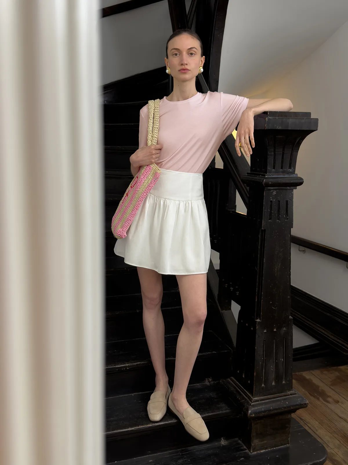 Model on black staircase wearing blush MARY T-Shirt, white skirt, beige loafers, and two-tone crocheted tote—Parisian street style