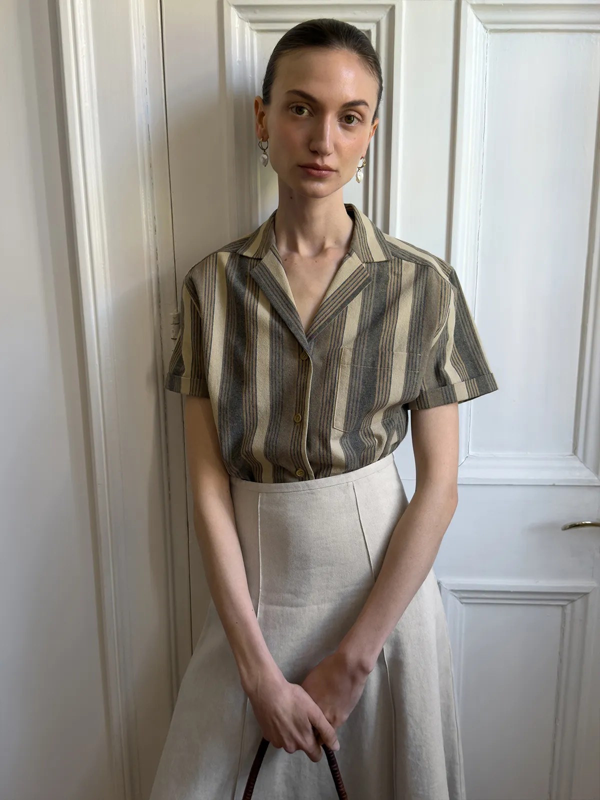 Model in REAGAN shirt with beige A-line skirt, hands clasped, against white door
