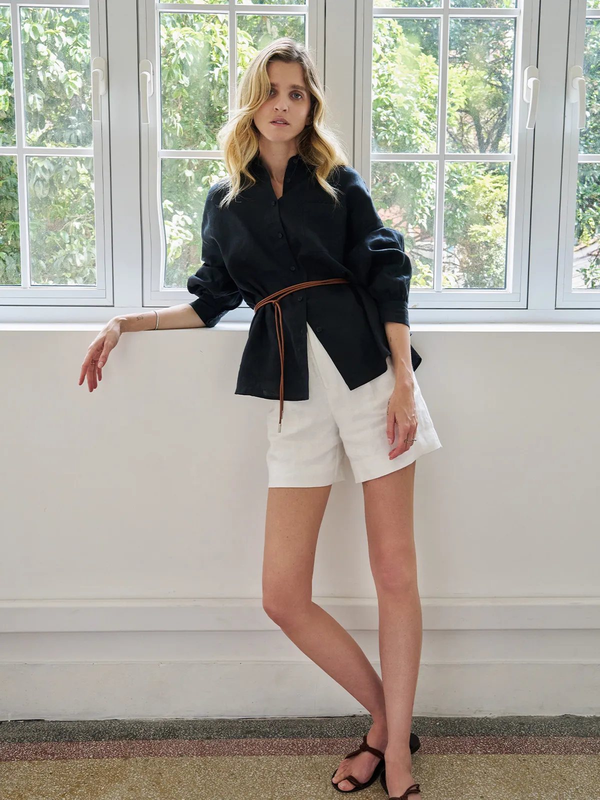 Model wearing black linen BREATH shirt with lantern sleeves, belted with leather cord, paired with white linen shorts in bright window light