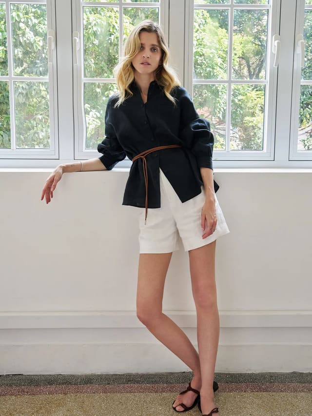 Model wearing black linen BREATH shirt with lantern sleeves, belted with leather cord, paired with white linen shorts in bright window light