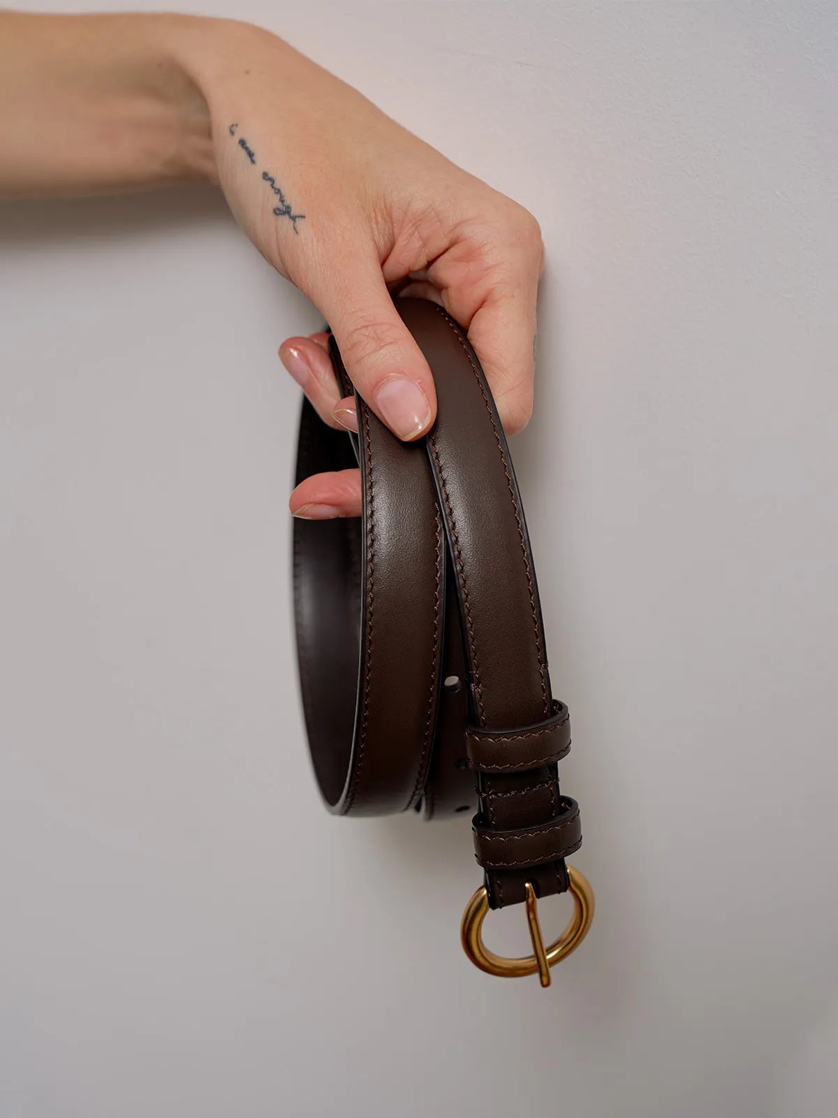 Hand holding chocolate brown KAY belt vertically against light gray wall, focusing on leather grain and buckle