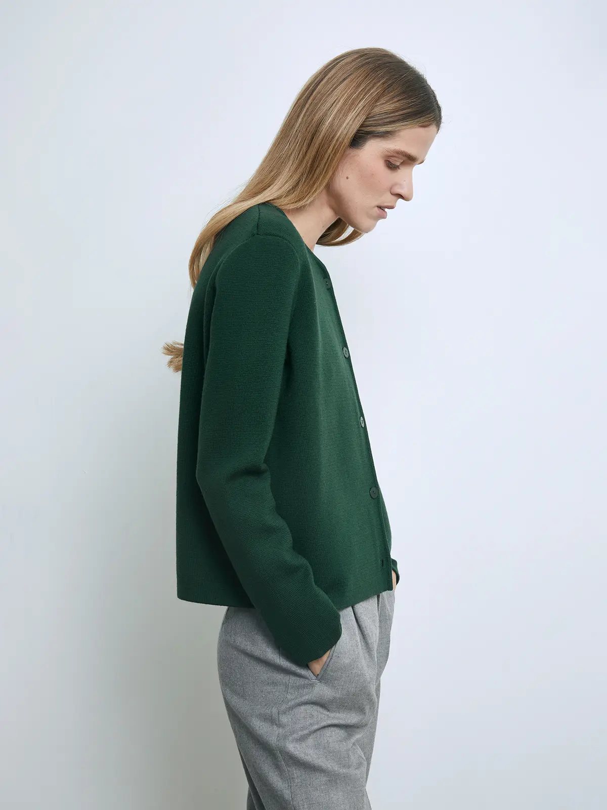 Side view of a model wearing a green knit cardigan with long sleeves and front buttons, paired with trousers.