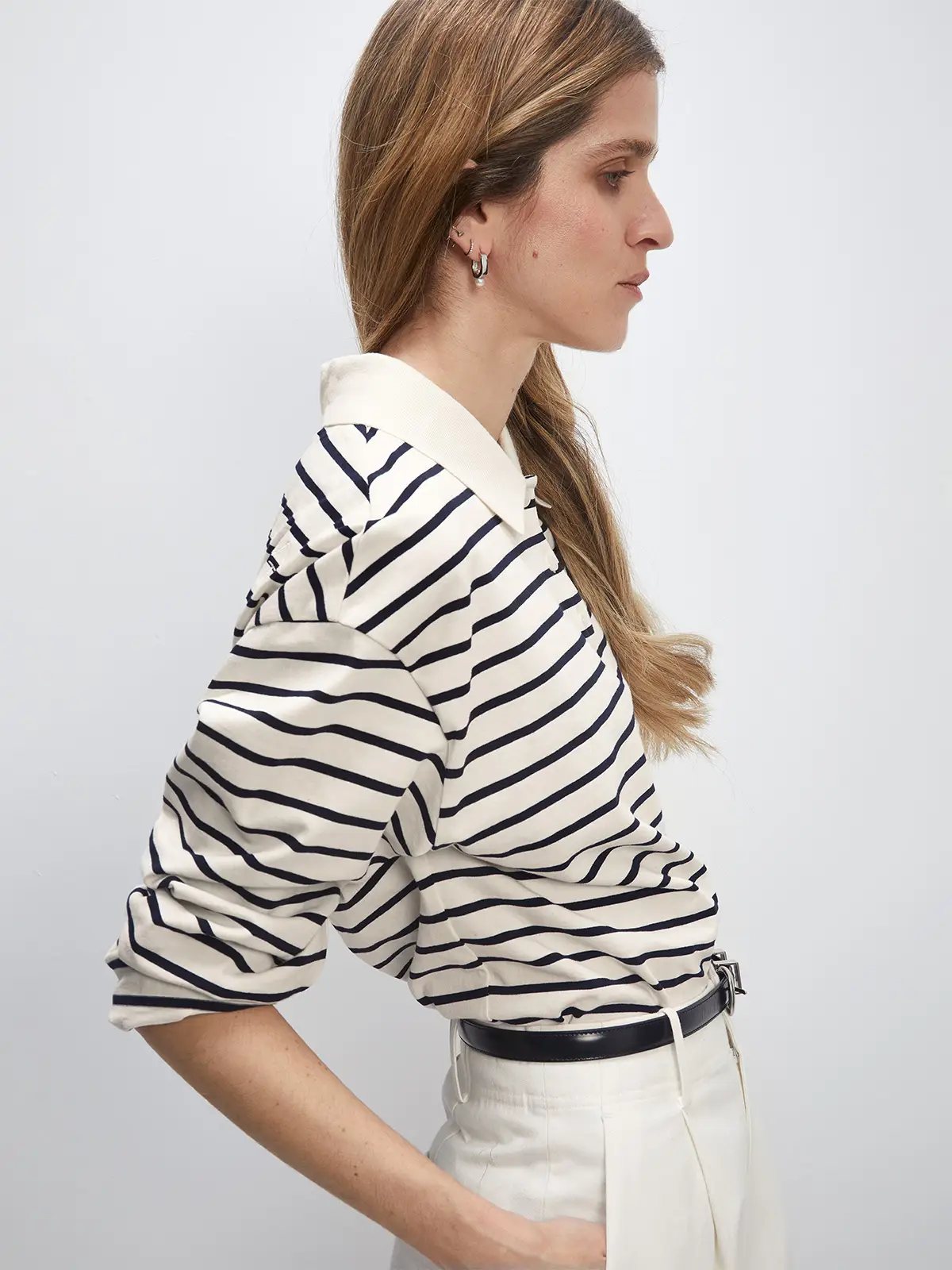 Side view of a model in a white/black striped long-sleeve polo collar cotton t-shirt with white trousers and a black belt.