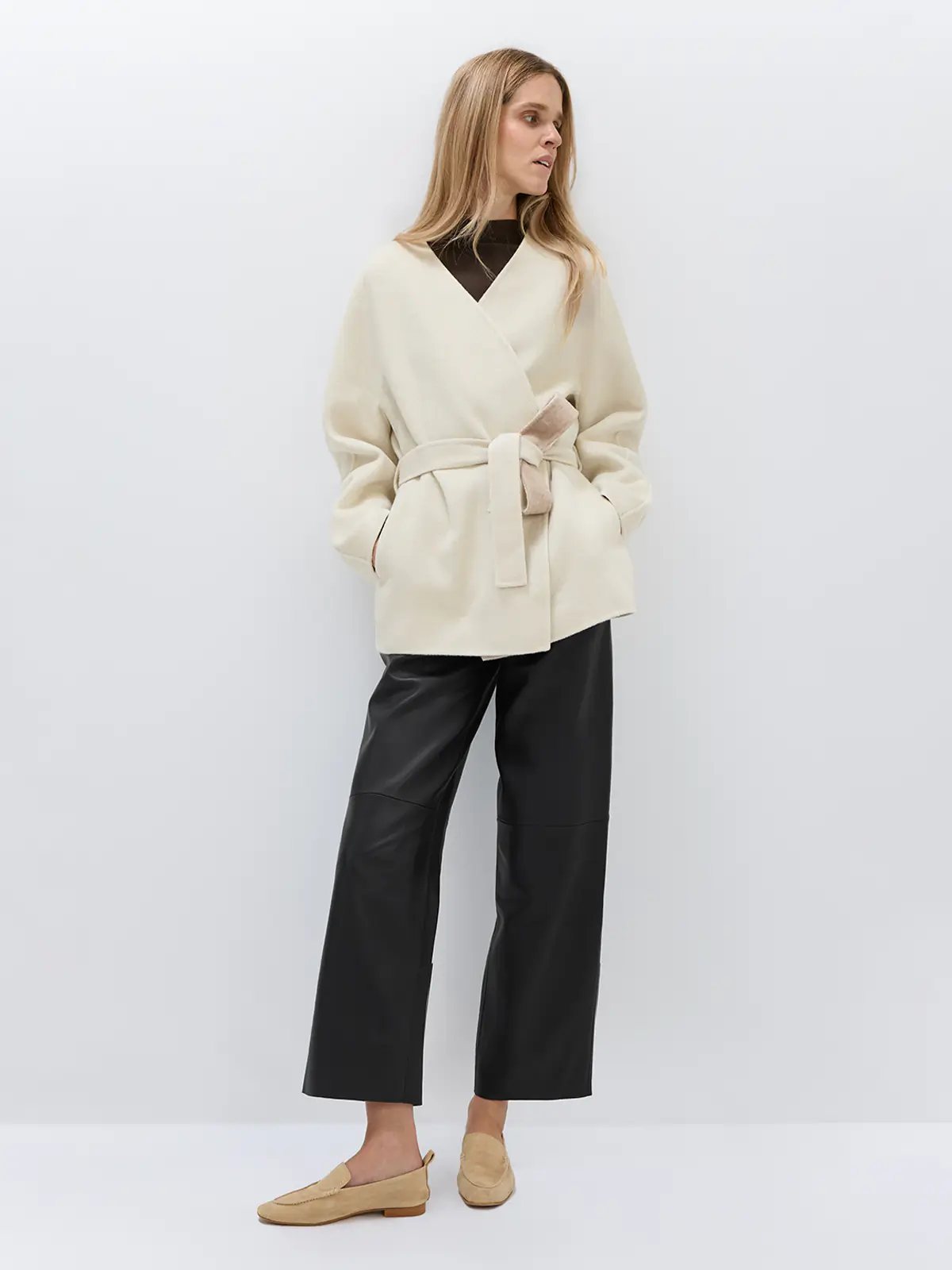 Model in an off-white wool lyocell belted coat with hands in pockets, showing wrap front and belt detail.