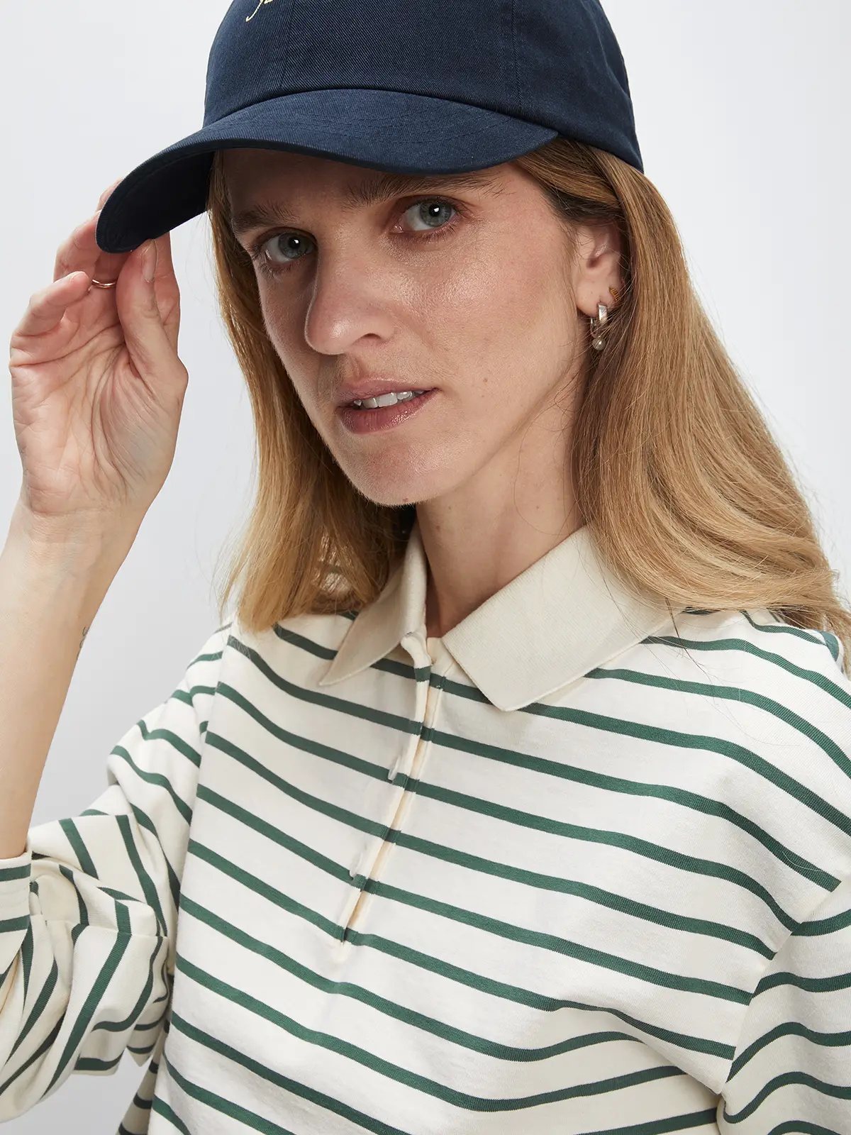 Model wearing a white/green striped long-sleeve polo collar cotton t-shirt with jeans and a cap, adjusting the brim.