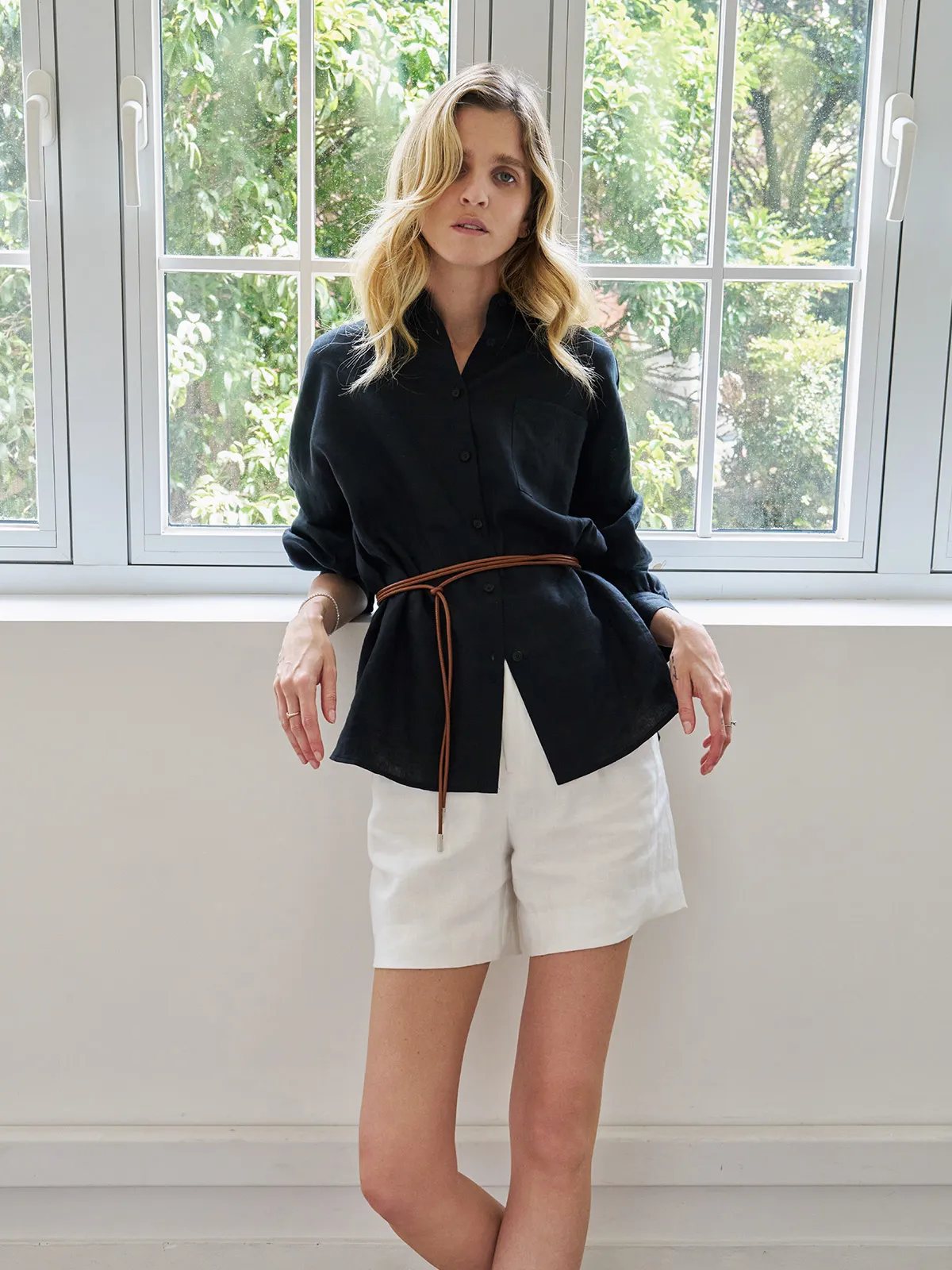 Front-facing model in black linen BREATH shirt with lantern sleeves, belted, paired with white shorts against white window frame