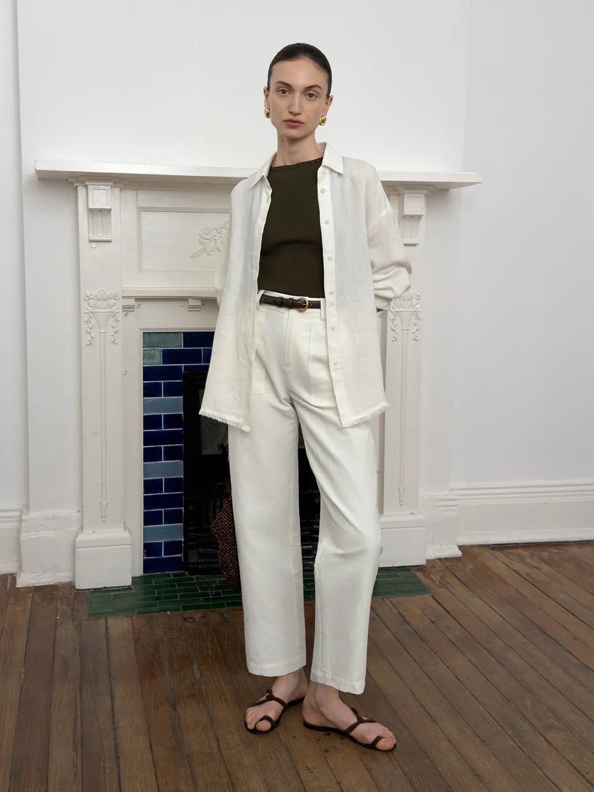 Model in white REID cropped trousers layered under open ivory shirt, olive ribbed tank, and brown belt against white fireplace