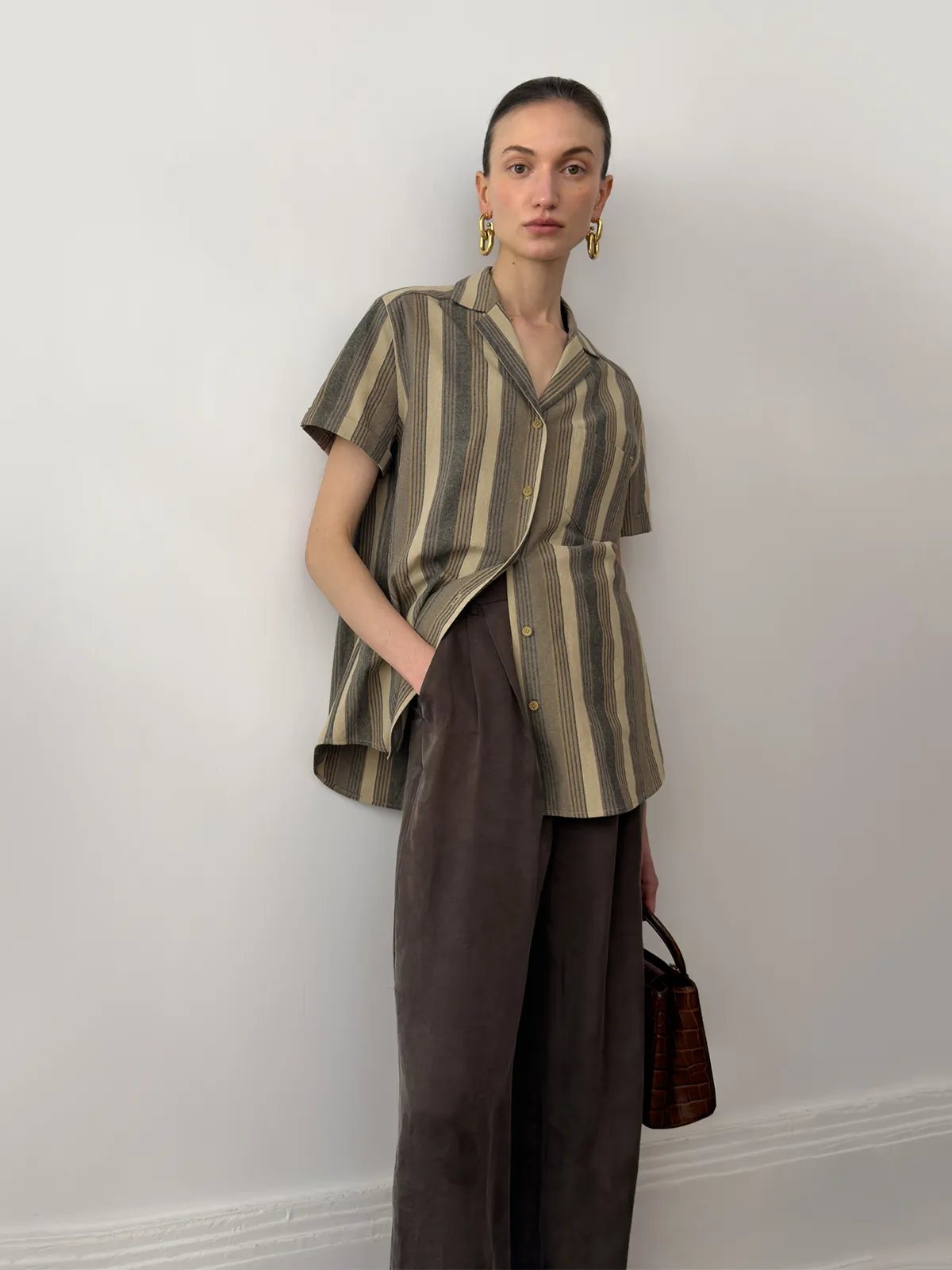 A model wearing a minimalist striped camp collar short-sleeve shirt paired with dark brown wide-leg trousers, representing a sophisticated quiet luxury aesthetic for an elevated everyday wardrobe.
