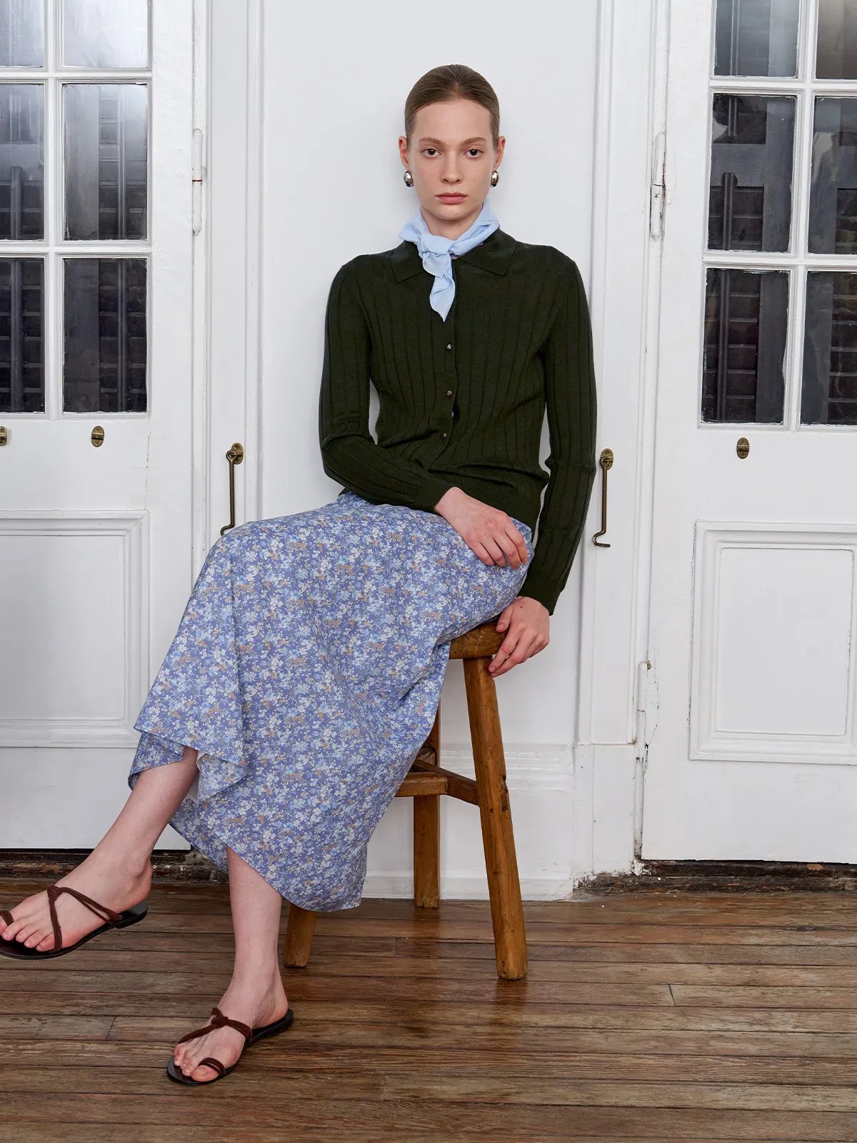 Model seated on wooden stool wearing olive green cardigan, floral skirt, and light blue TIANA scarf tied at neck