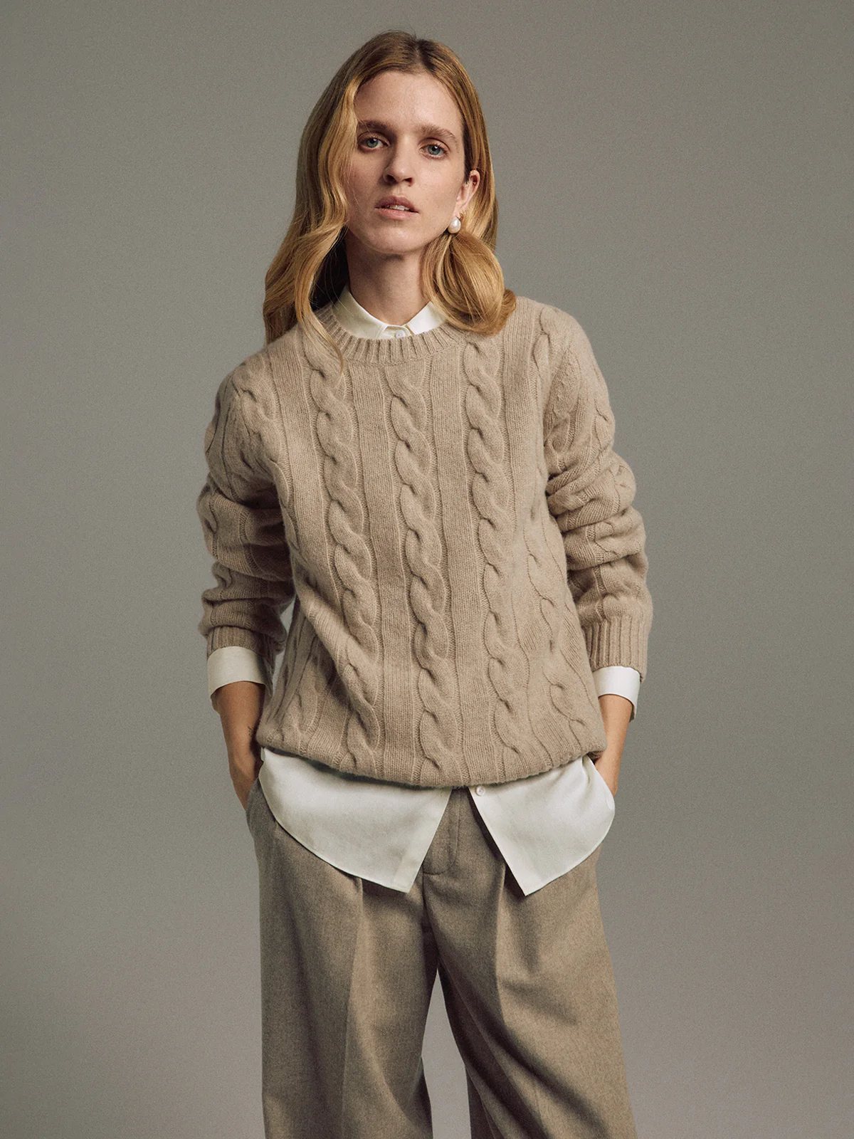 Model wearing an apricot cable knit jumper layered over a shirt, paired with wide-leg trousers and hands in pockets.