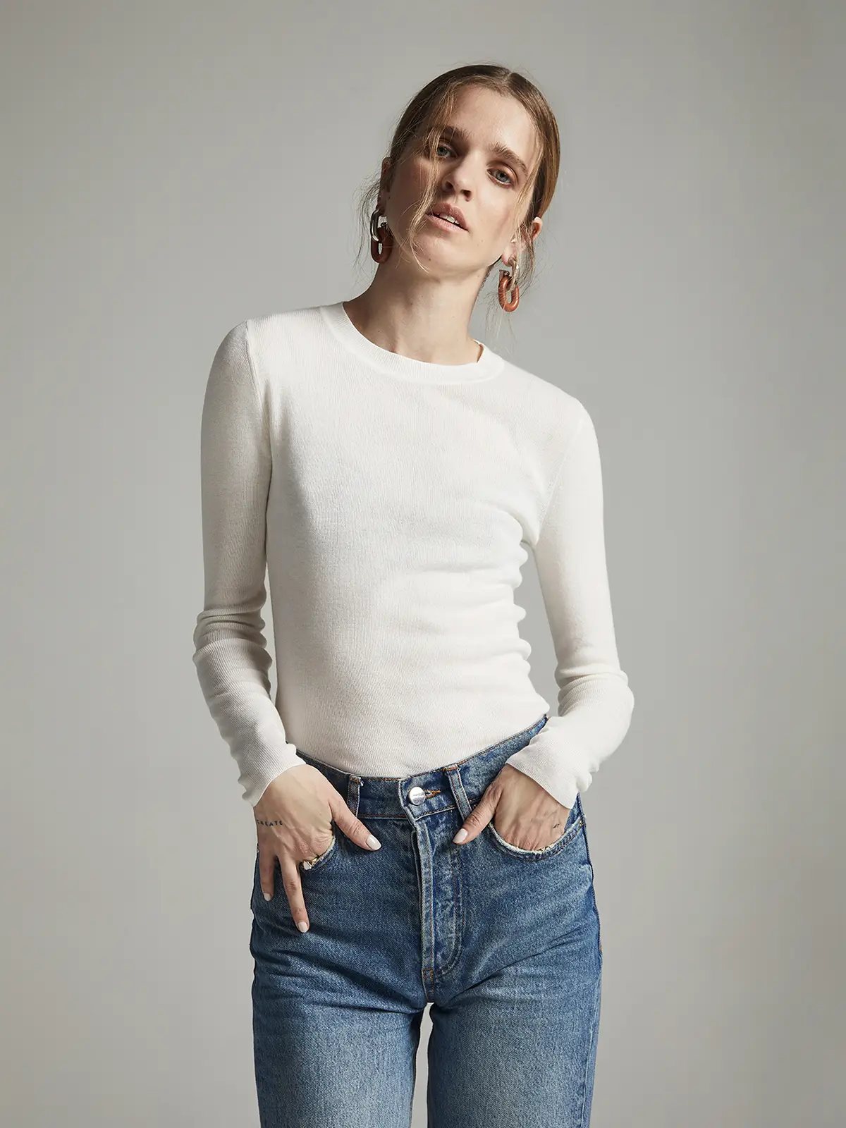Model wearing a slim-fit, long-sleeve knit top in White with hands in denim pockets.