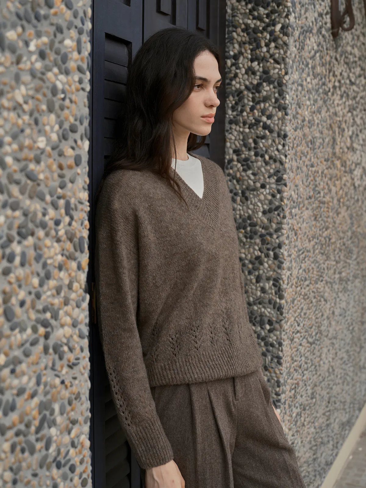 Model in taupe AIRY Jumper layered over white tee, paired with matching taupe trousers, against pebble wall