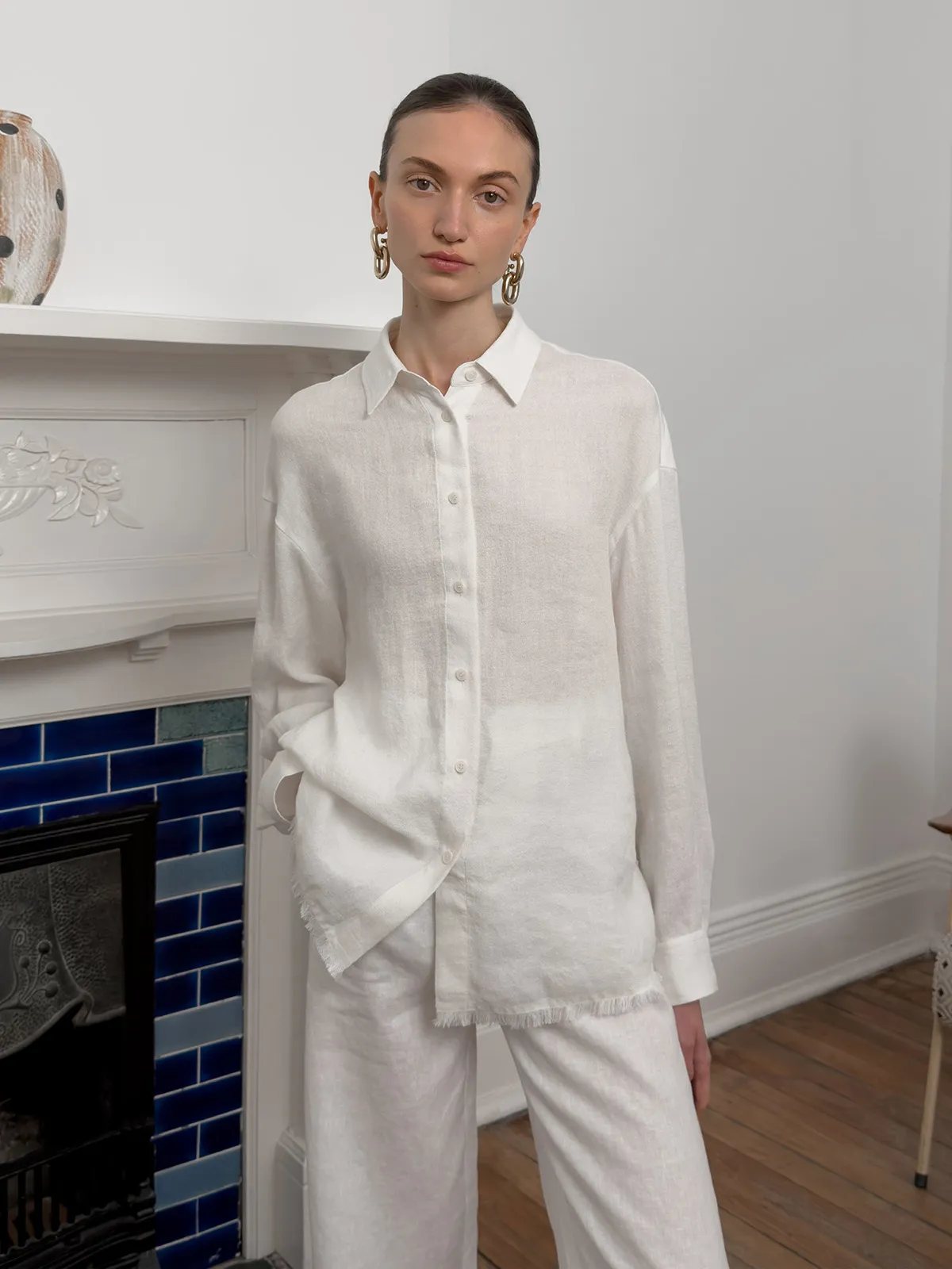 Front-facing model in white ELARA shirt and trousers beside blue-tiled fireplace, minimalist interior styling