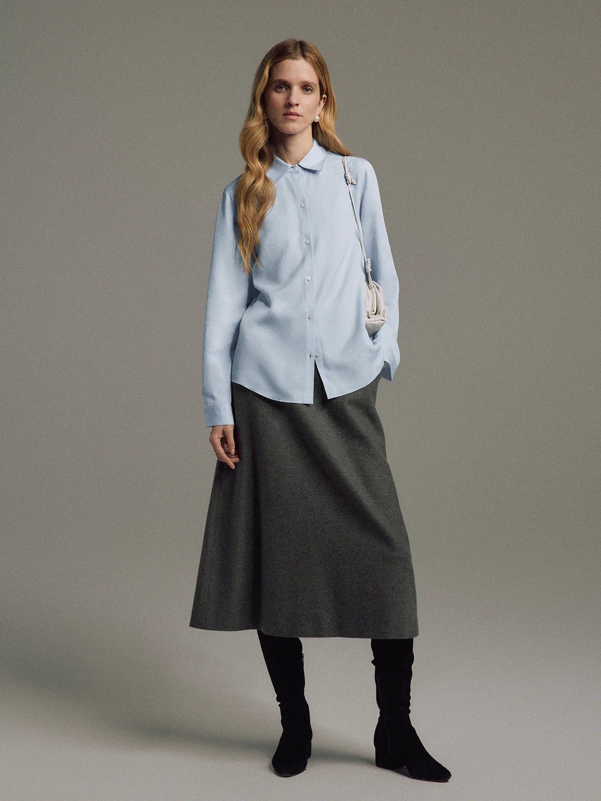 Full front view of a model in a Gray wool-blend A-line midi skirt, light shirt, boots, and a small shoulder bag.
