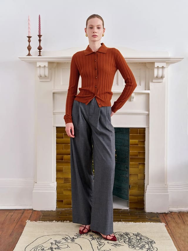 Model wearing terracotta CONSTANCE cardigan with grey pinstripe trousers in front of white fireplace