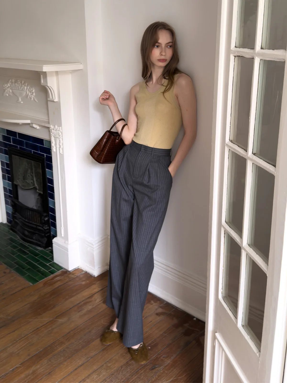 Model leaning against wall in ORAN trousers with beige tank, brown croc-effect bag, and suede flats