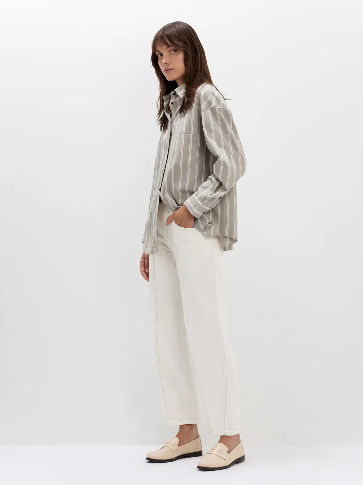 Side view of model wearing white jeans with contrast stitching, a striped button-up shirt, and holding a white handbag.