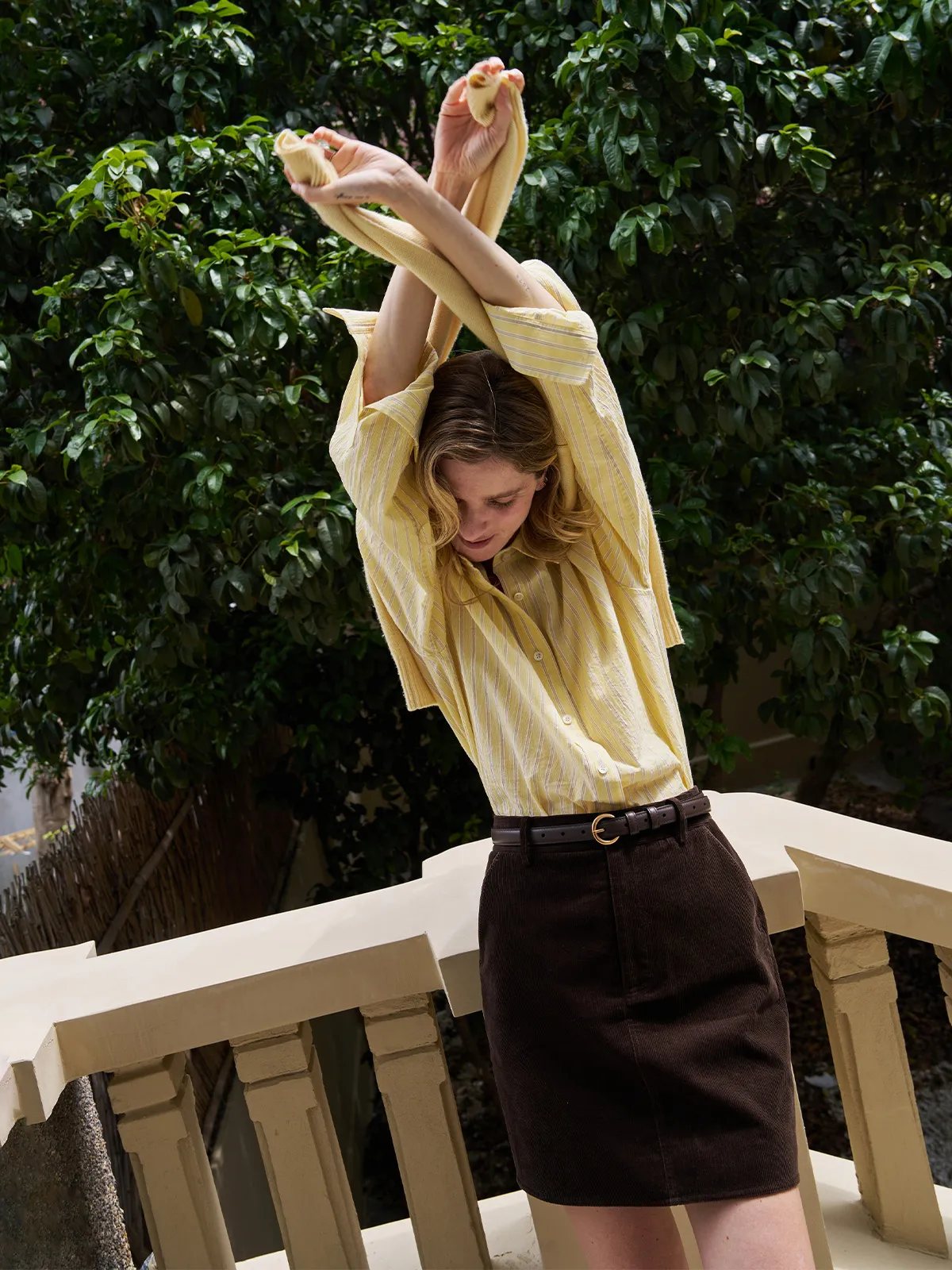 Model stretching arms overhead in yellow striped shirt and DENVER brown corduroy skirt on balcony