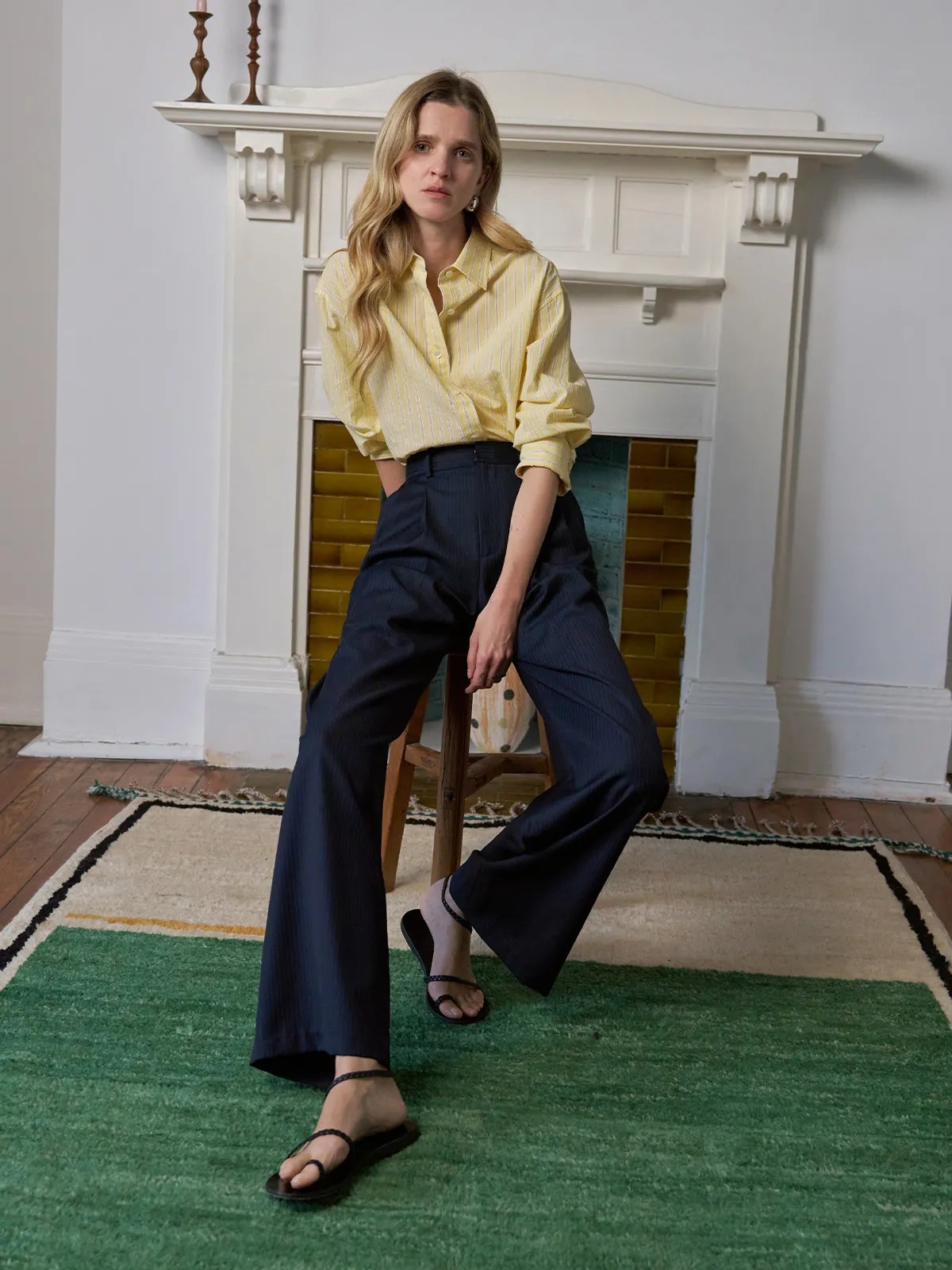 Model wearing ROGAN wide leg trousers with yellow striped shirt, seated on stool in vintage room setting