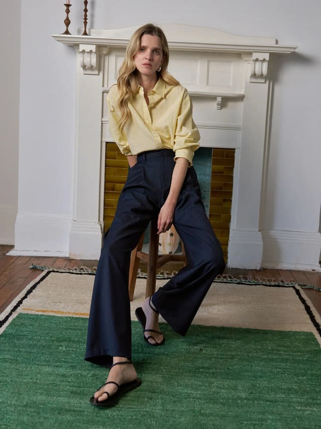 Model wearing ROGAN wide leg trousers with yellow striped shirt, seated on stool in vintage room setting