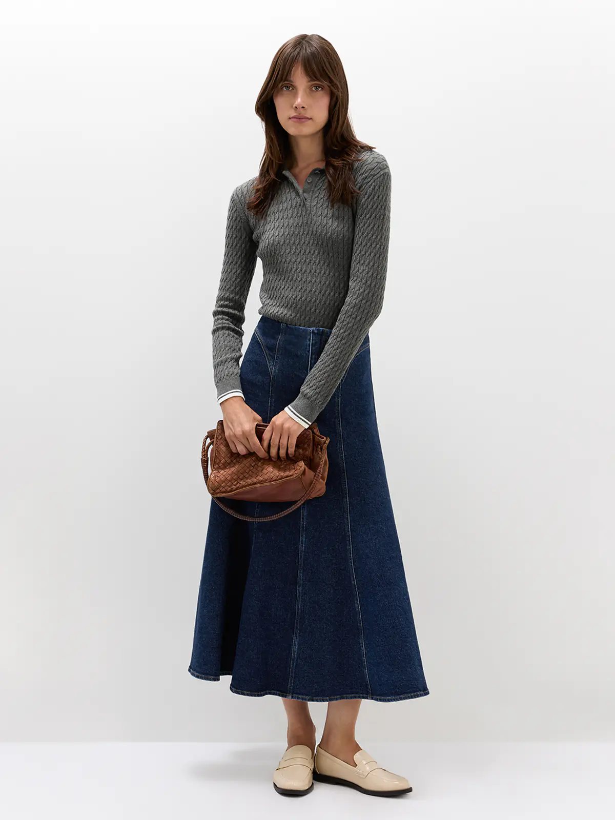 Side view of model wearing a Grey cable knit polo with long sleeves, ribbed cuffs, and a collar, paired with a skirt.