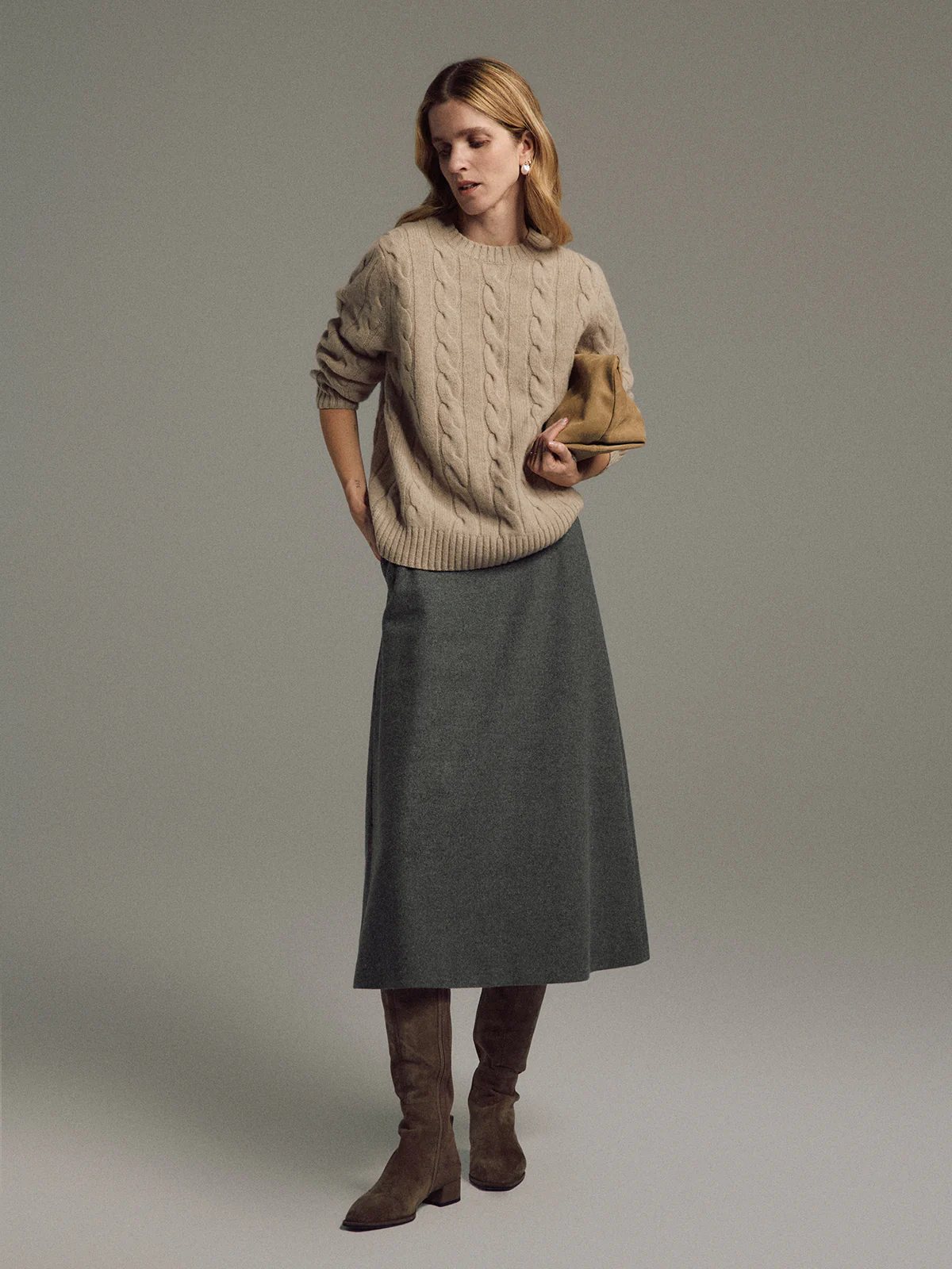 Side view of a model wearing a Gray wool-blend A-line midi skirt, cable-knit sweater, boots, holding a clutch.