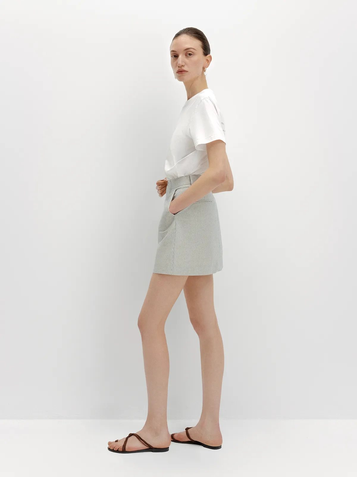Side profile of andreatang DEVON Mini Skirt with white tee and leather thong sandals