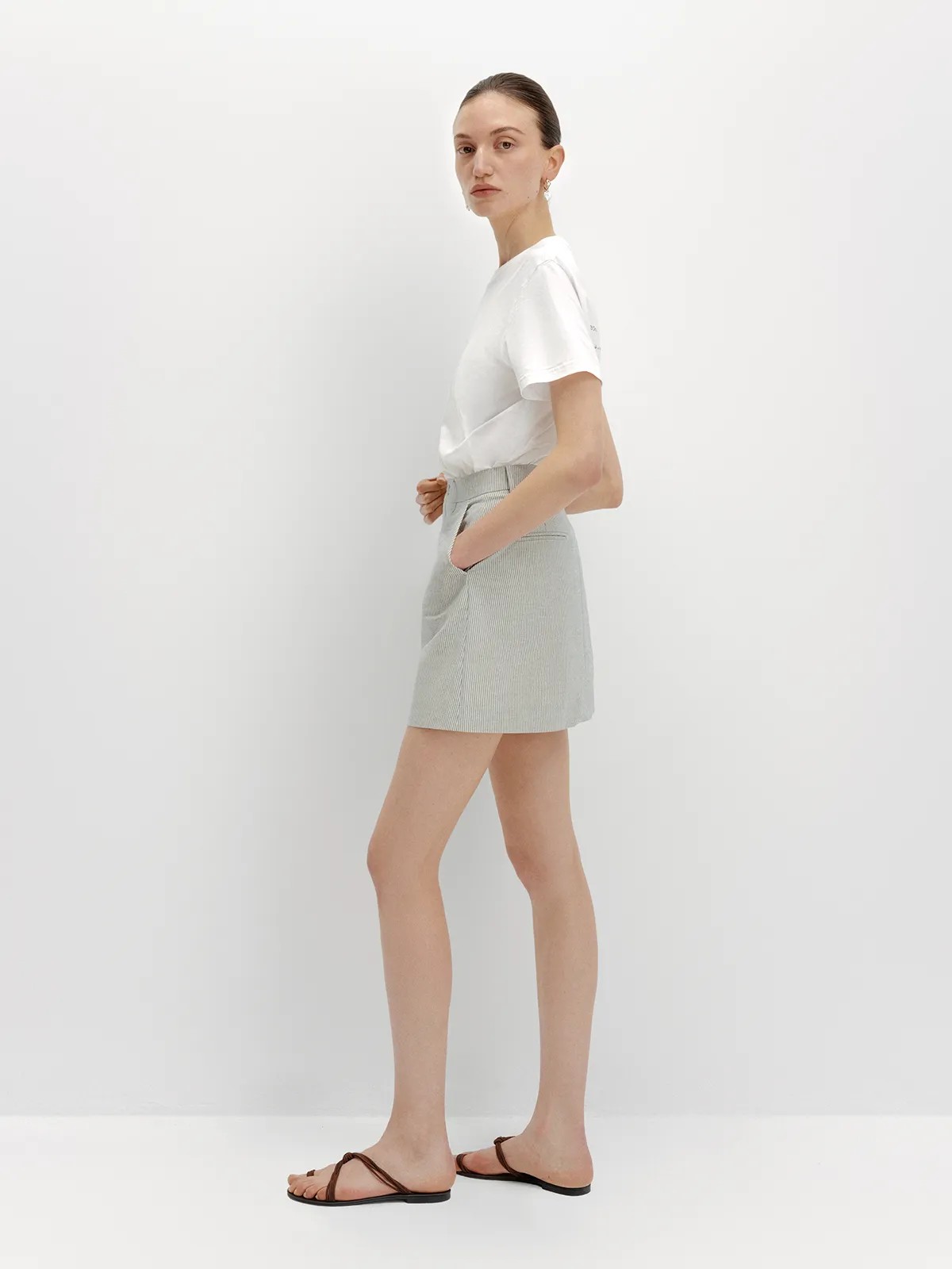 Side profile of andreatang DEVON Mini Skirt with white tee and leather thong sandals