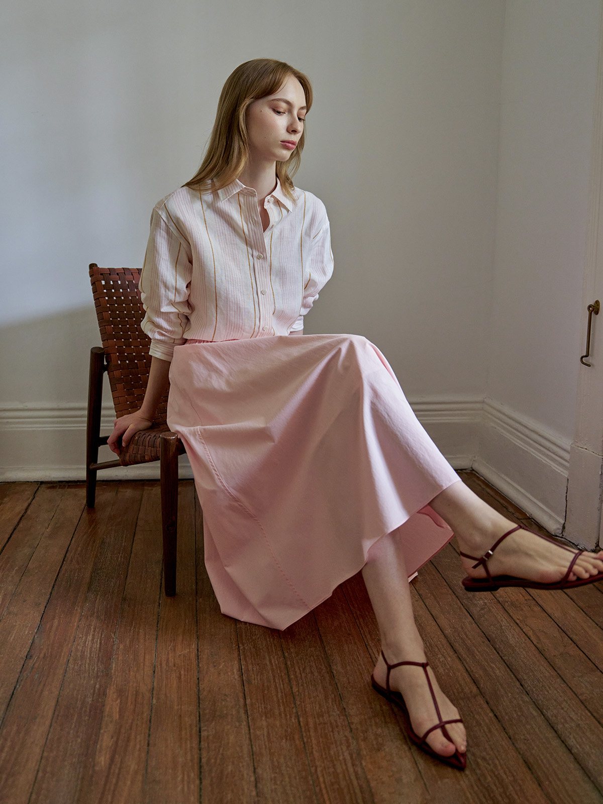 Blush maxi skirt with striped shirt, seated on wicker chair, side-angle composition