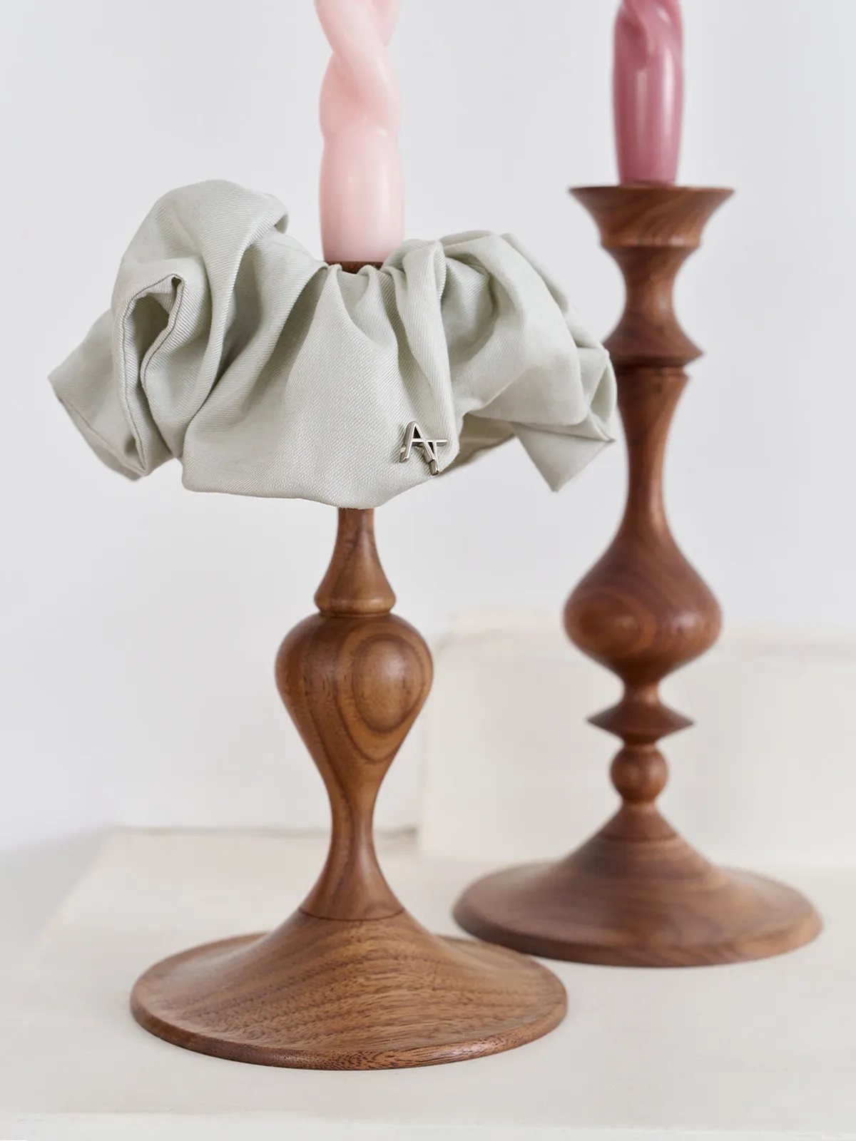 Sage green silk scrunchie draped over wooden candlestick with pink twisted candle, minimalist still life