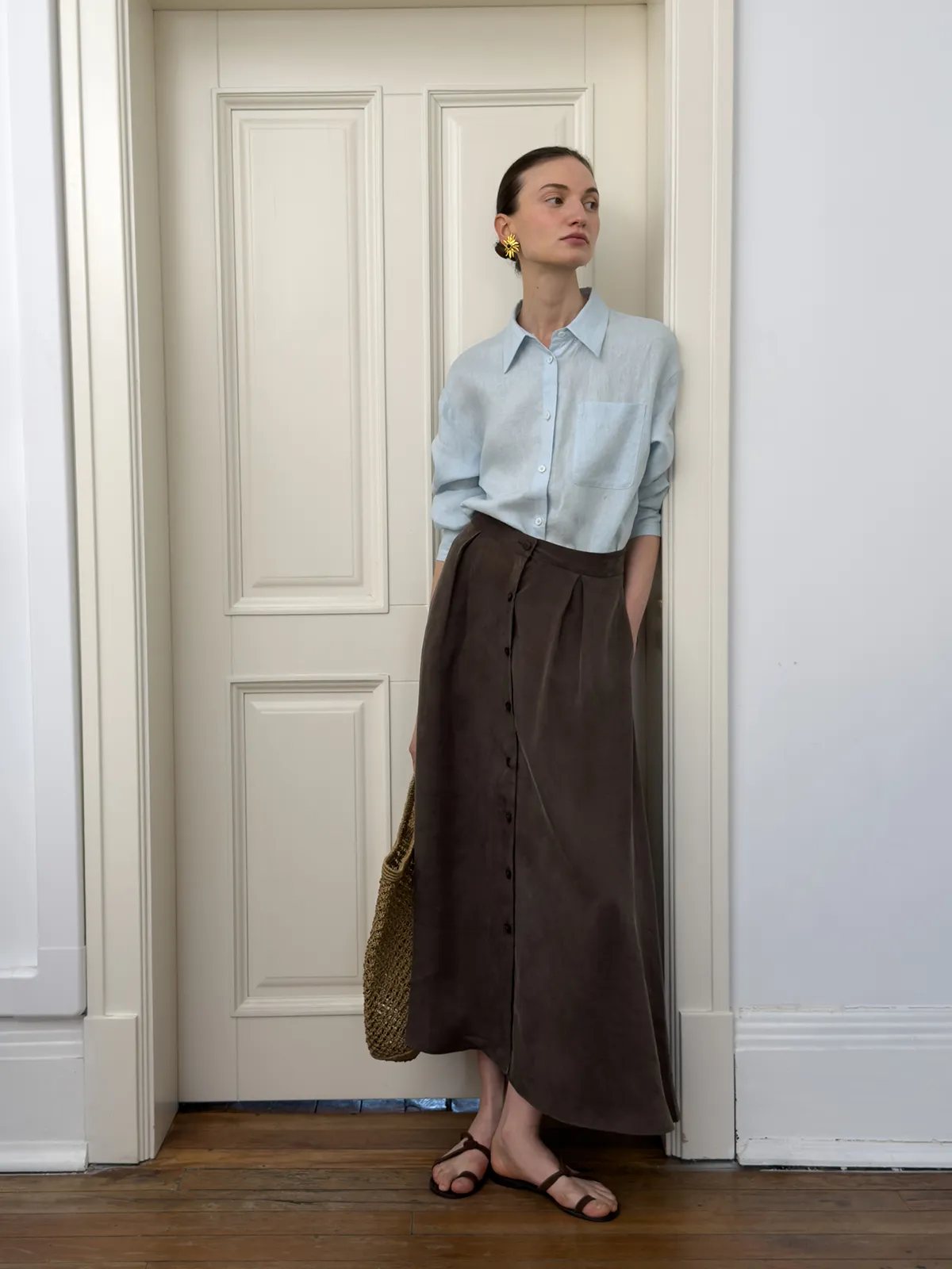 Brown NOBU Midi Skirt with light blue linen shirt, woven tote, and gold earrings