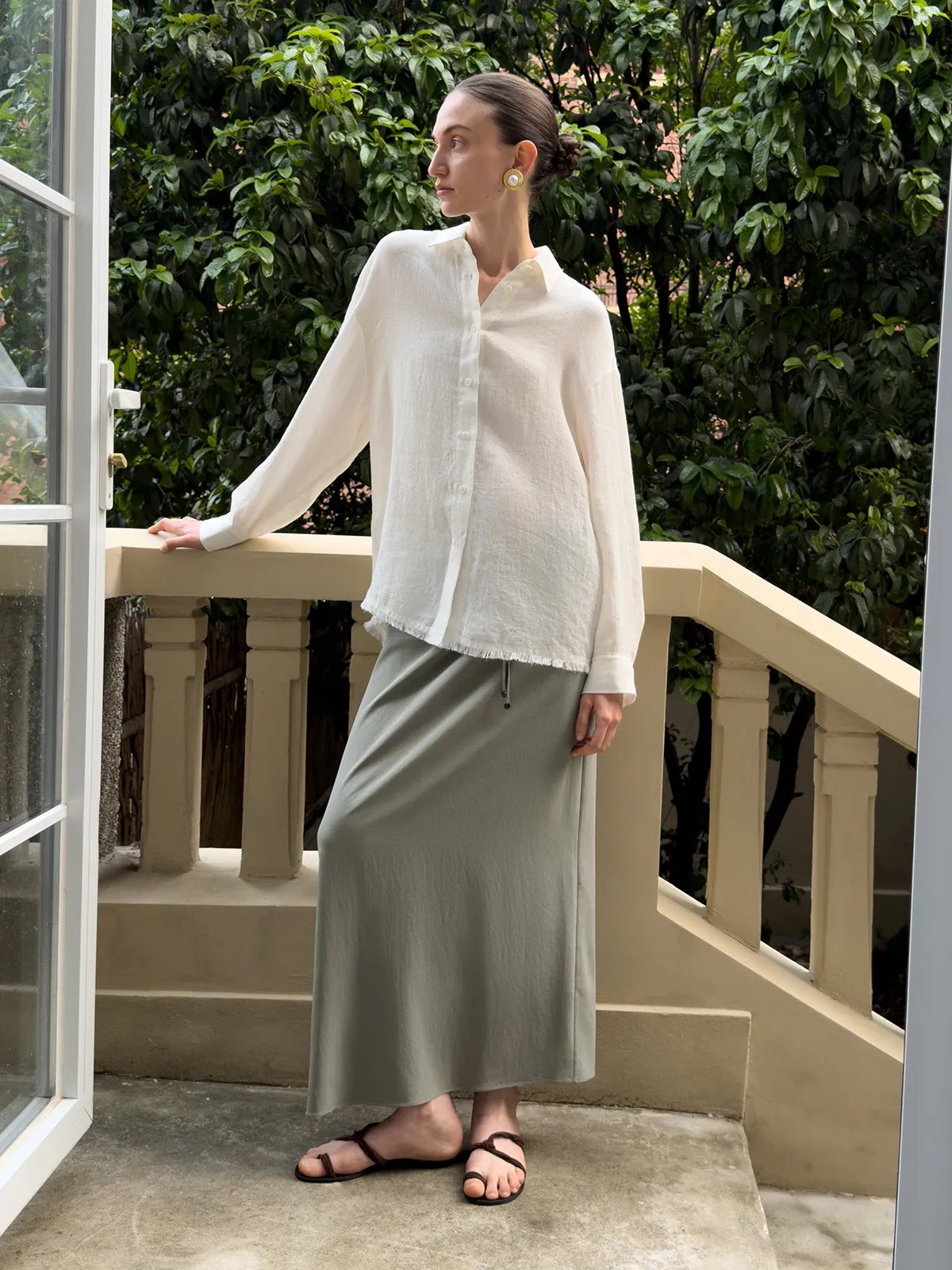 Model wearing ELARA shirt with olive green maxi skirt on sunlit balcony, lush greenery background