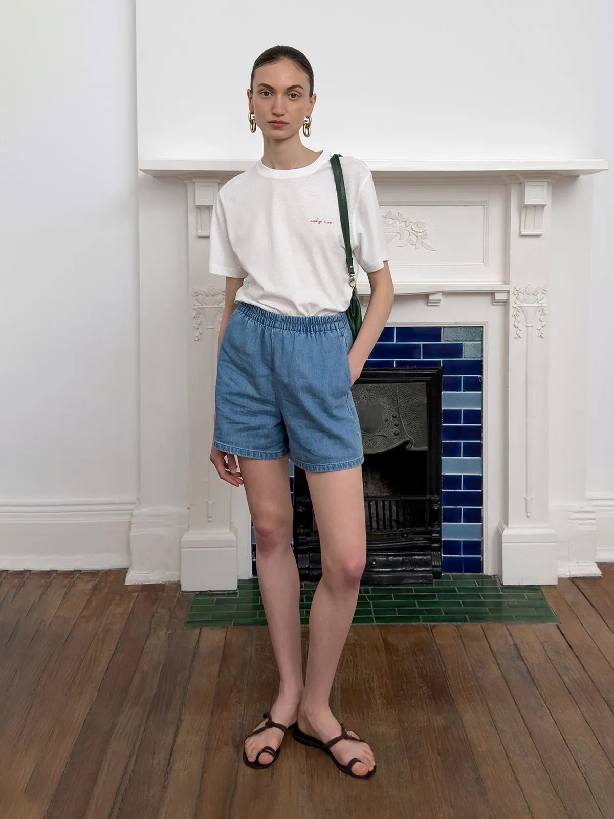 Model in white relaxed tee with blue denim shorts and brown sandals, standing before ornate fireplace