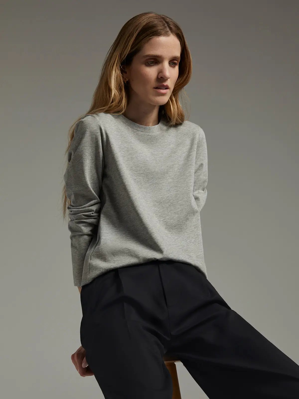 Model wearing a grey long-sleeve crewneck T-shirt with rolled sleeves, seated on a stool with dark trousers.