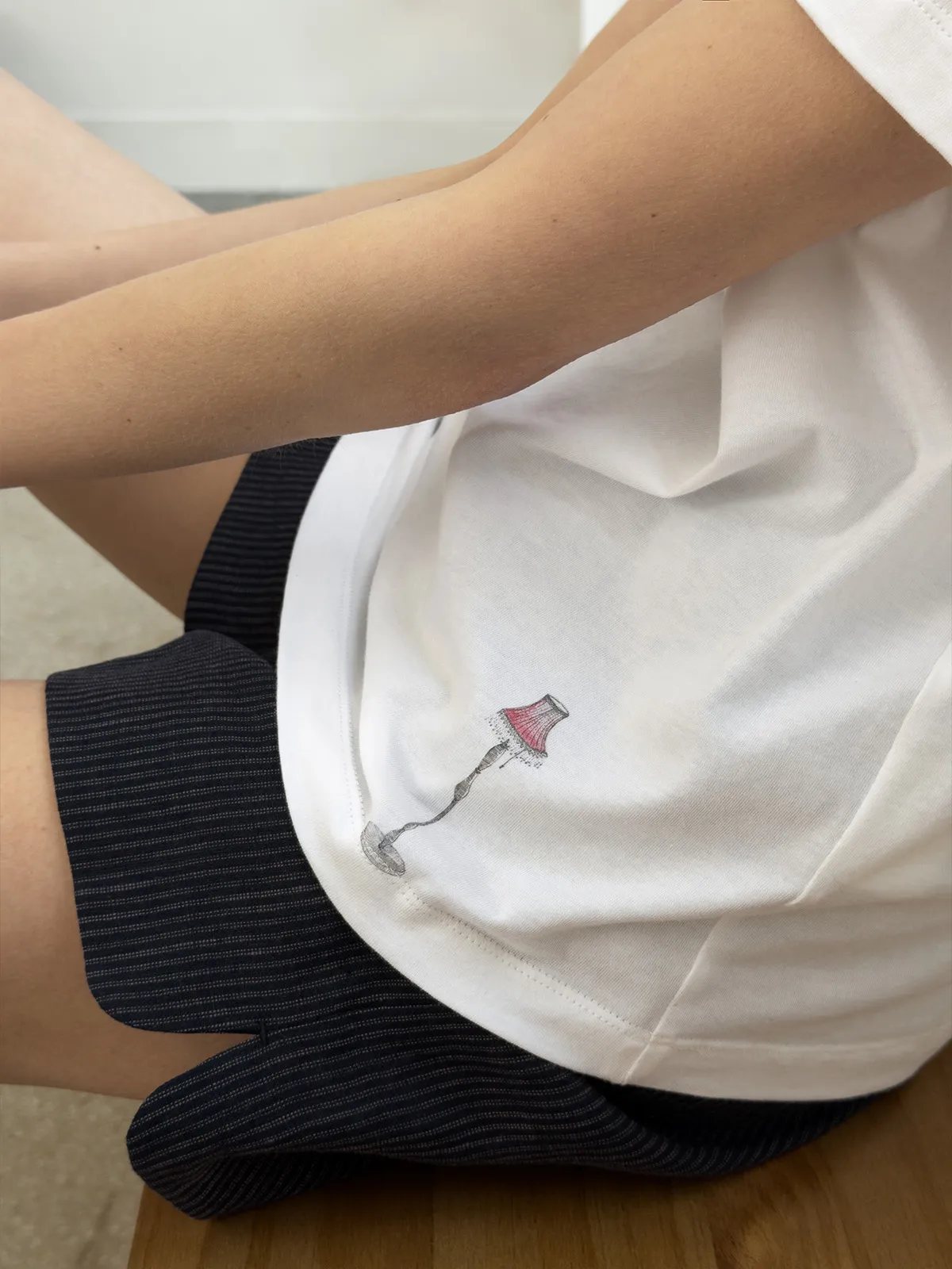 Hidden detail: miniature embroidered table lamp illustration on lower right hem of EDWARD white t-shirt — pink shade, silver base, quiet luxury storytelling