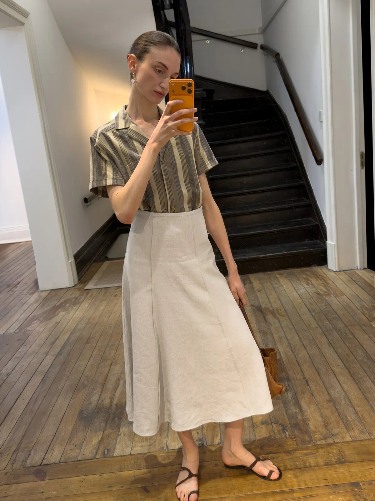 Mirror selfie: model in striped shirt and NATASHA skirt holding orange phone, staircase background