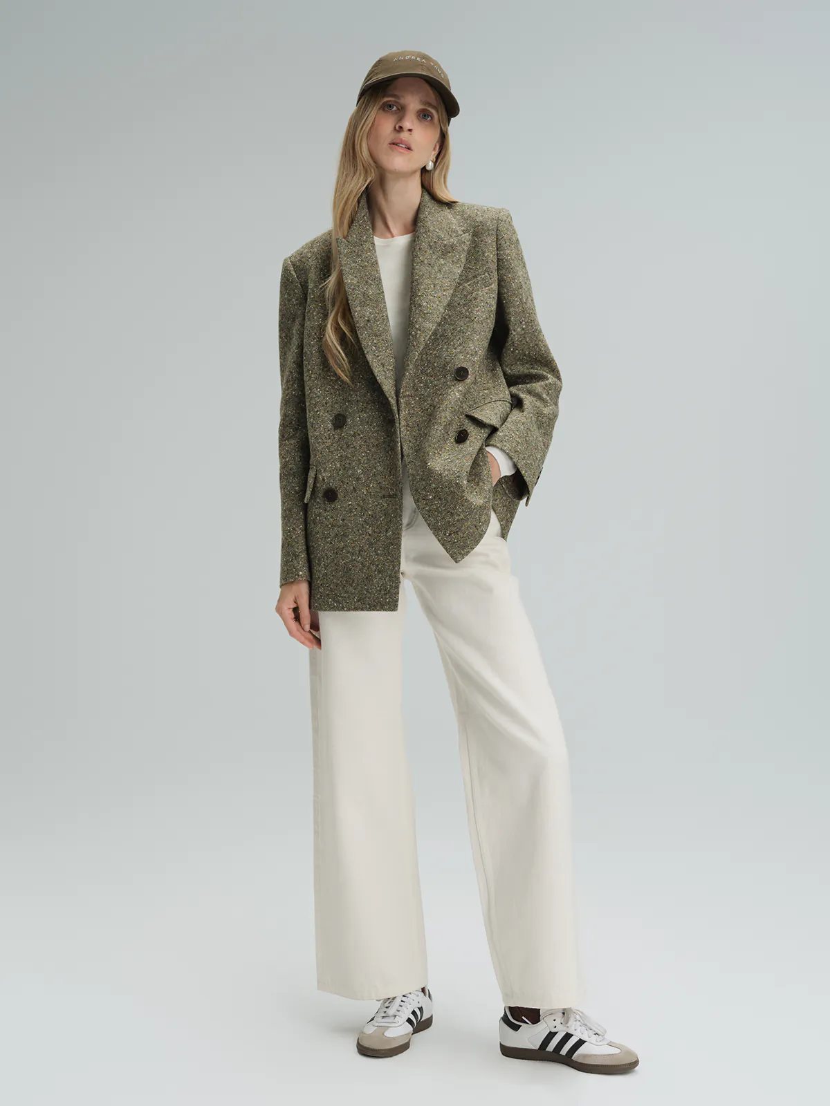 Front-facing model wearing a green wool flecked tweed double-breasted blazer over a turtleneck, holding a bag.