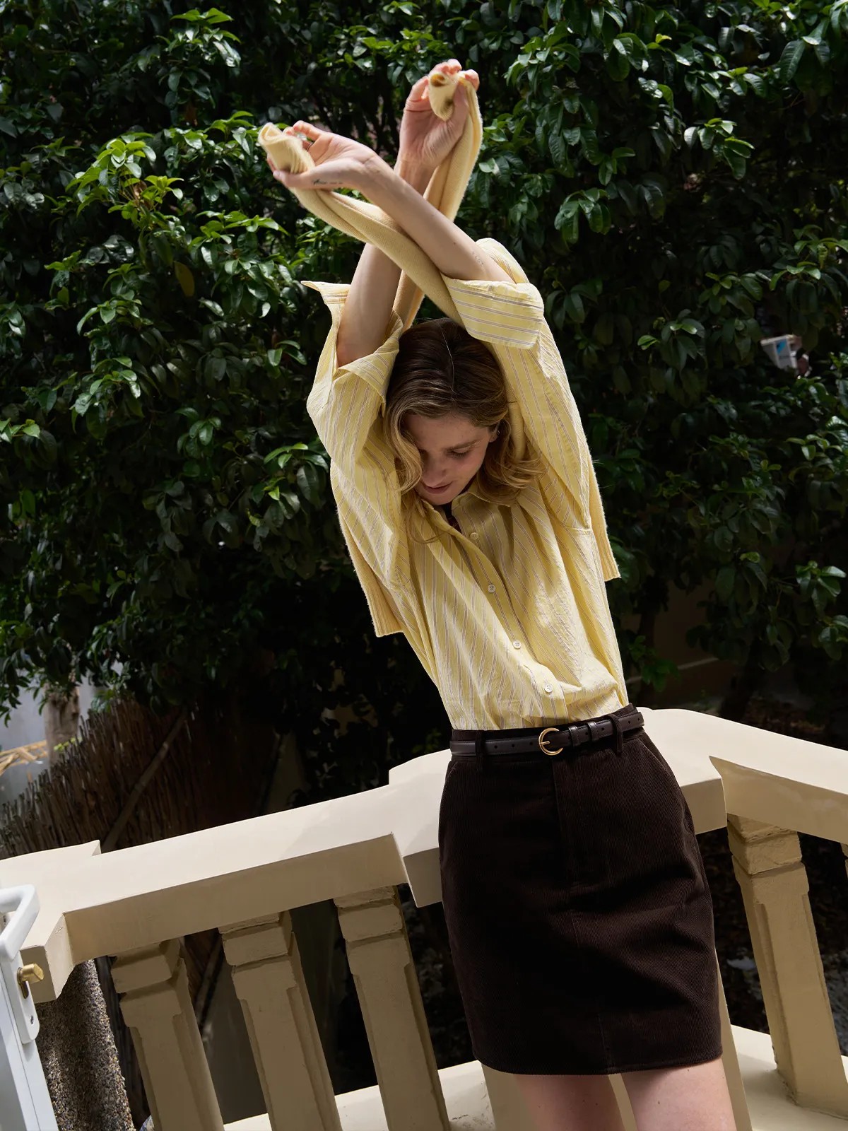 Model stretching arms overhead, wearing yellow striped shirt, brown corduroy skirt, and chocolate KAY belt