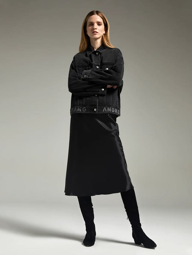 Model wearing andreatang washed-black vintage denim jacket with logo hem, styled with black satin midi skirt and knee boots