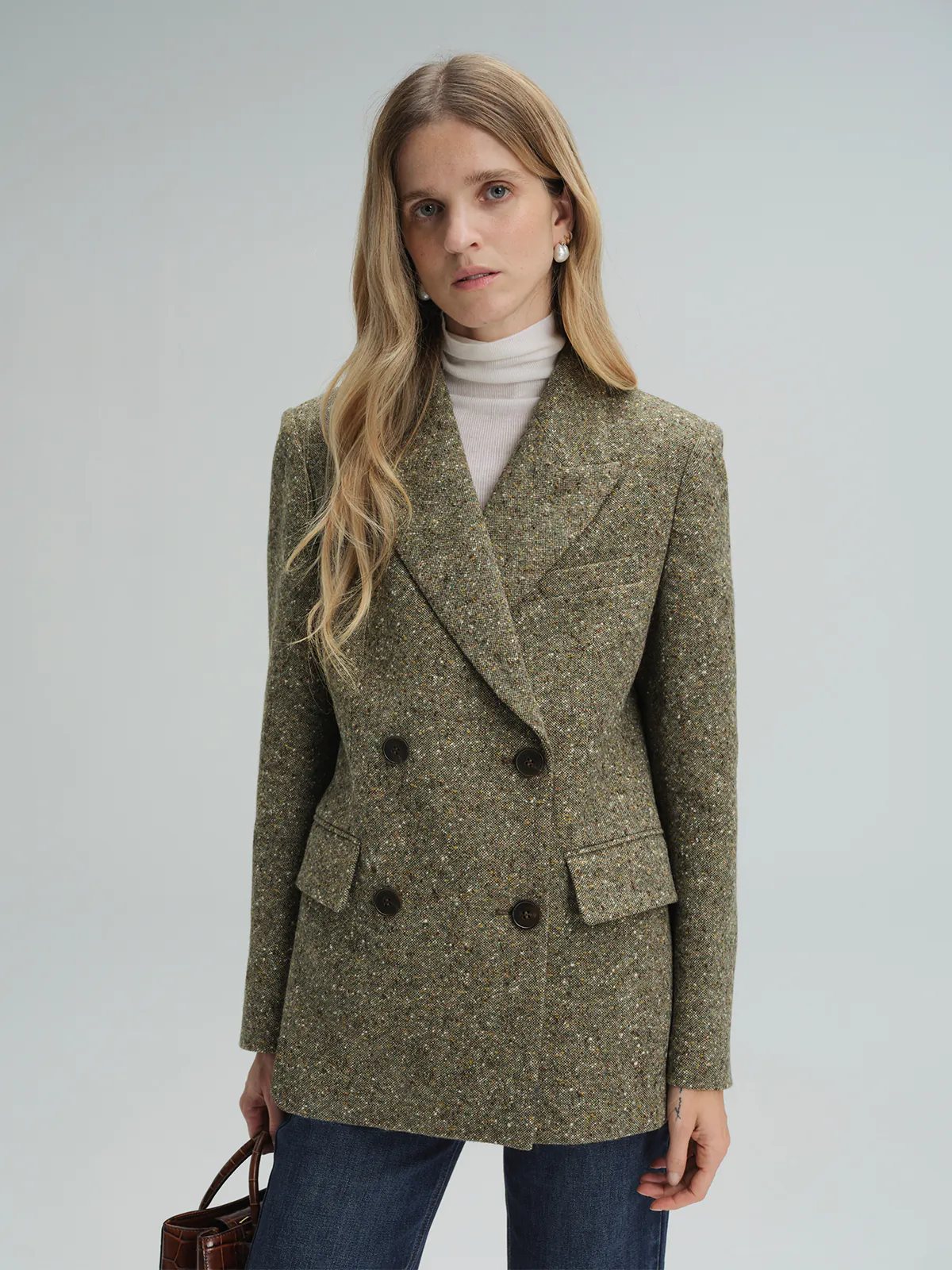 Front-three-quarter view of a model in a green wool flecked tweed double-breasted blazer, hands in pockets, styled with a blouse and dark trousers.